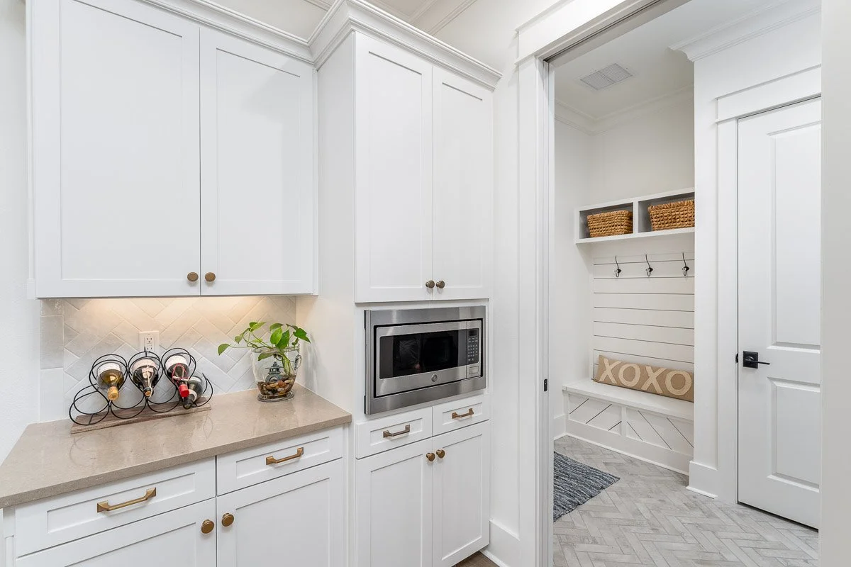Real Estate photo of a white kitchen with cabinets, countertop, and built-in microwave; looking into a mudroom with hooks, cushions, and baskets.