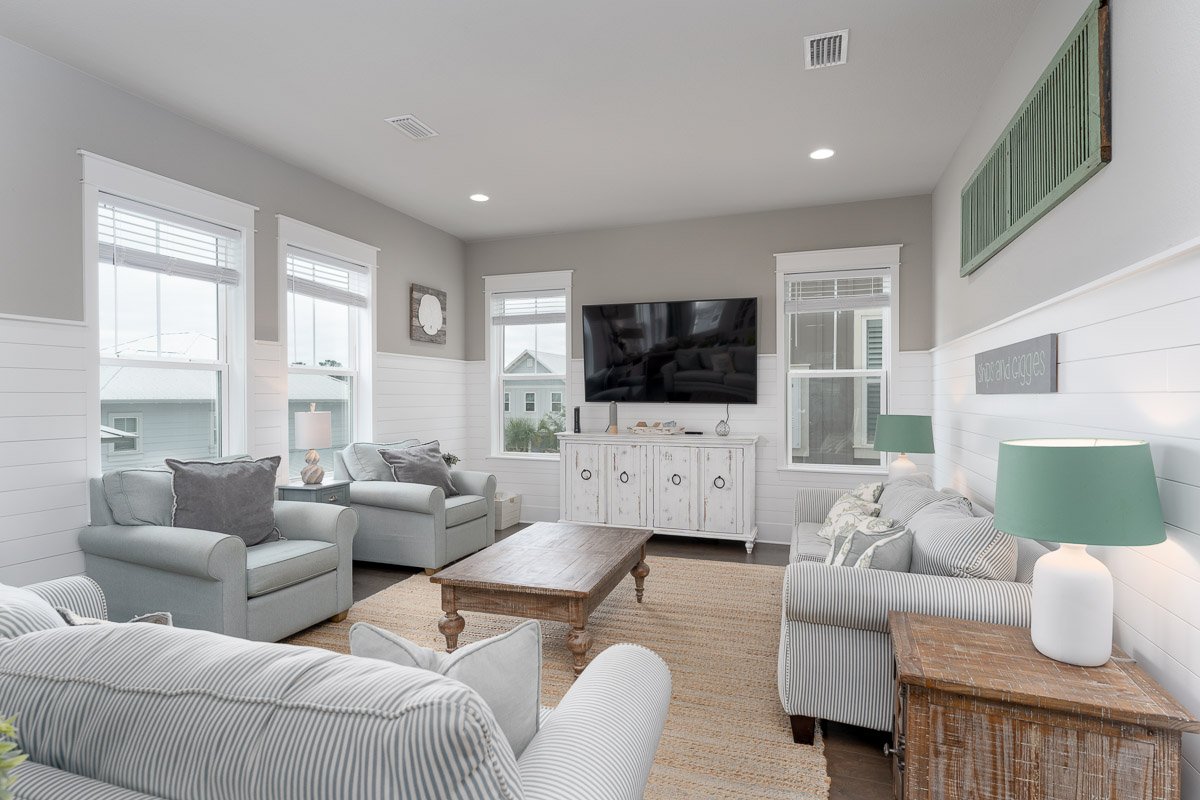 Living room with white and gray striped sofas, mint green lamps, a wooden coffee table, a white distressed cabinet, a wall-mounted TV, and windows with white blinds.