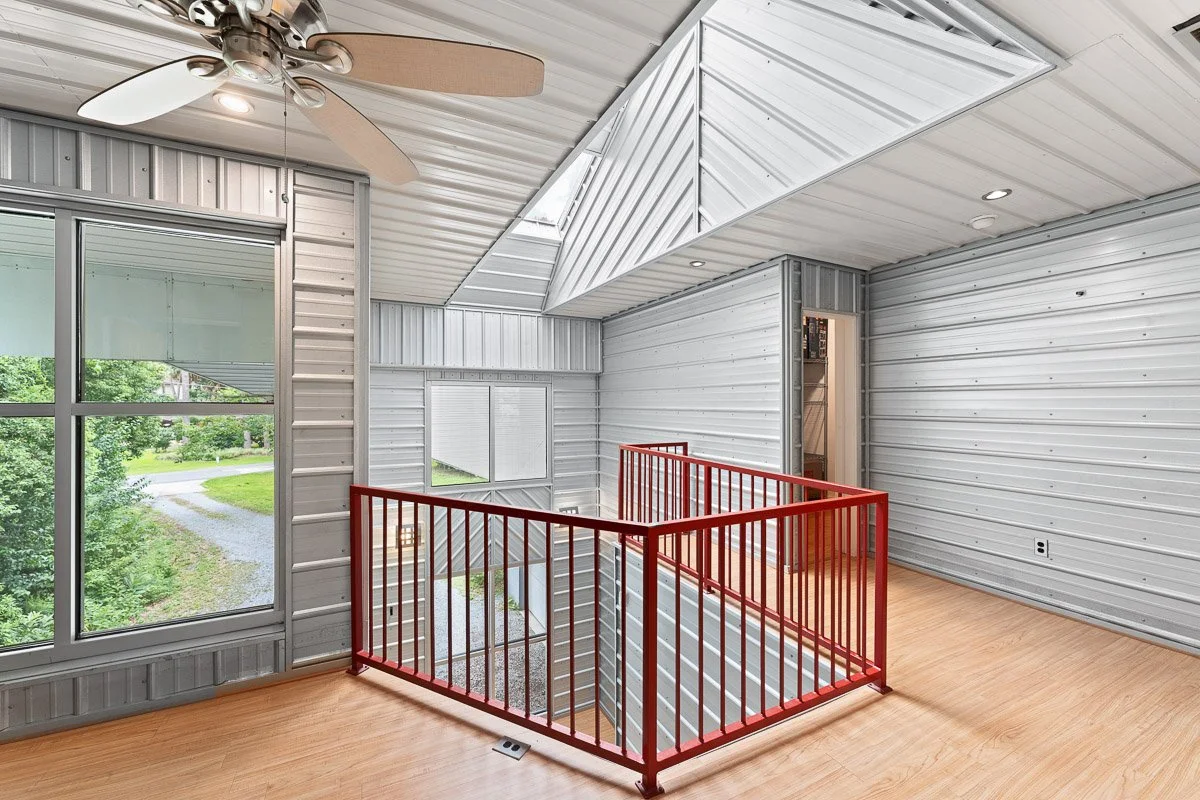 Real Estate photo of a modern loft with metal siding walls, large windows, a ceiling fan, and a wooden floor, overlooking the staircase with a red safety railing.
