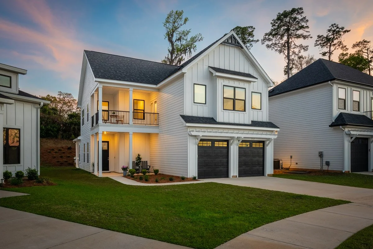 Real Estate photo of a modern two-story white house with black garage doors and a front porch during sunset.
