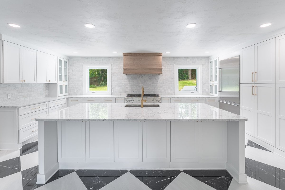 Real Estate photo of a bright, newly renovated, modern kitchen with white cabinets and a large central island with a marble countertop, two windows behind the stove, and black and white patterned floor tiles.