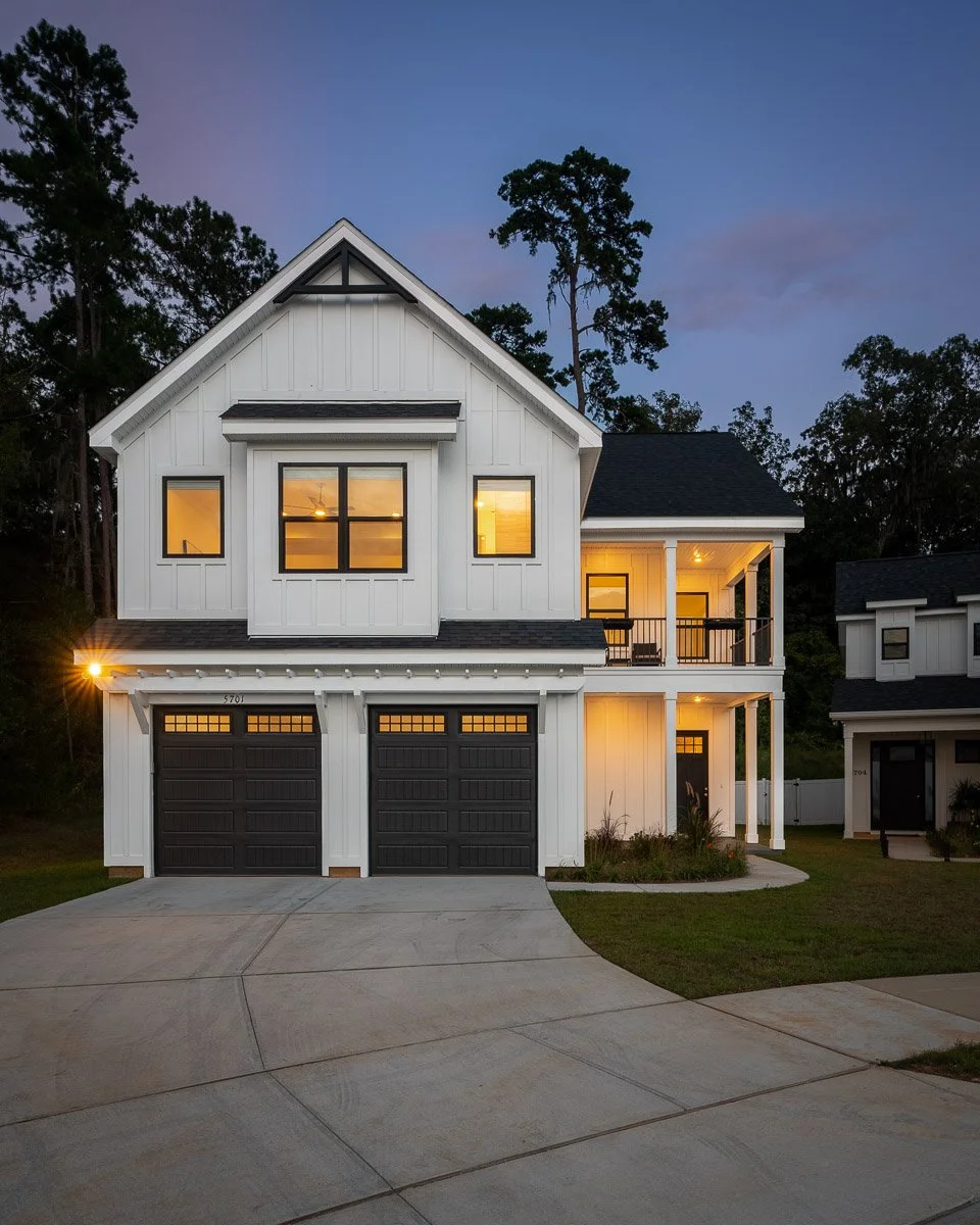 Real Estate photo of a modern two-story house with a white exterior, black garage doors, and warm interior lighting, situated at dusk with trees in the background.