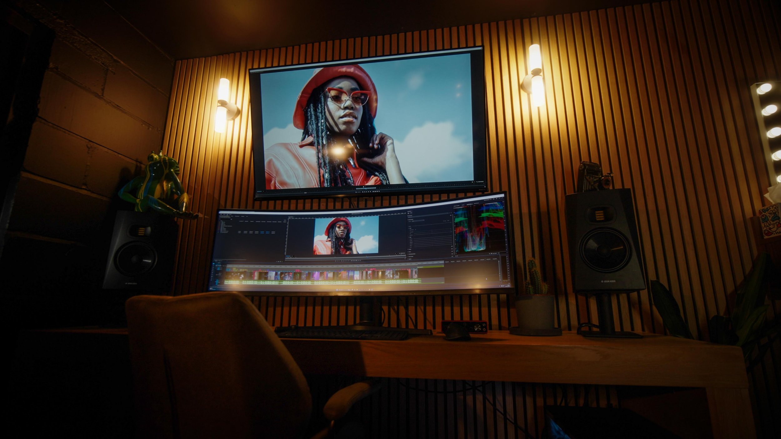 A professional video editing setup with two monitors displaying a woman in a red hat and sunglasses, surrounded by studio speakers and decorative items in a room with wooden panel walls.
