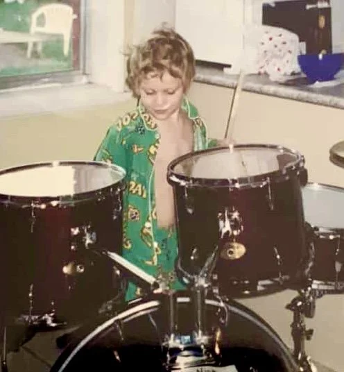 A young boy with curly hair wearing green pajamas, standing behind a drum set in a kitchen.