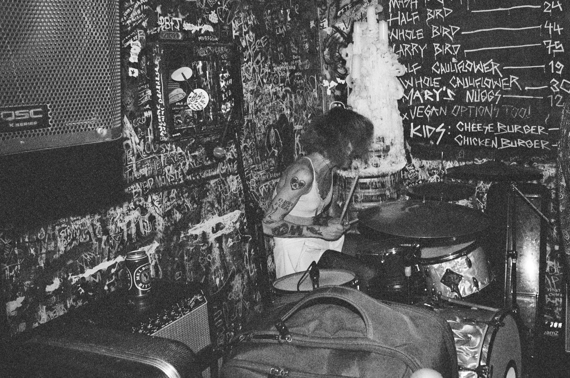 A person with tattoos on their arm is playing drums in a room filled with graffiti and chalkboard menus with food options and prices.