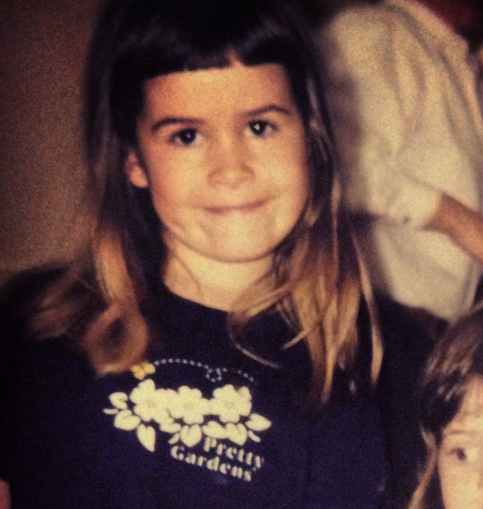 A young girl with long light brown hair and a slight smile, wearing a black shirt with white floral embroidery and the text 'Sapporo Gardens'.