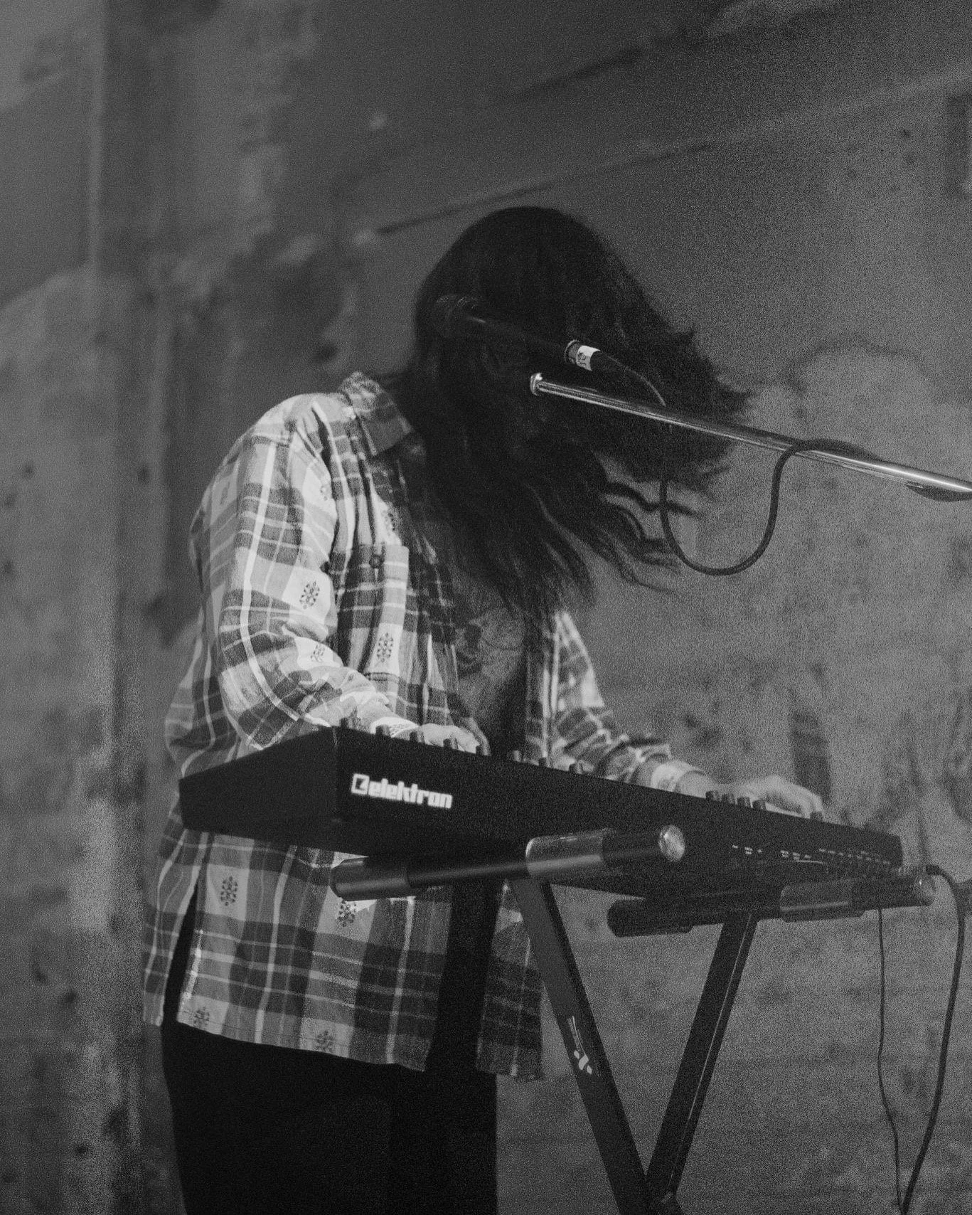 A person with long hair wearing a plaid shirt, playing a keyboard with the brand name 'Keebtron' in a dimly lit room with a brick wall background.