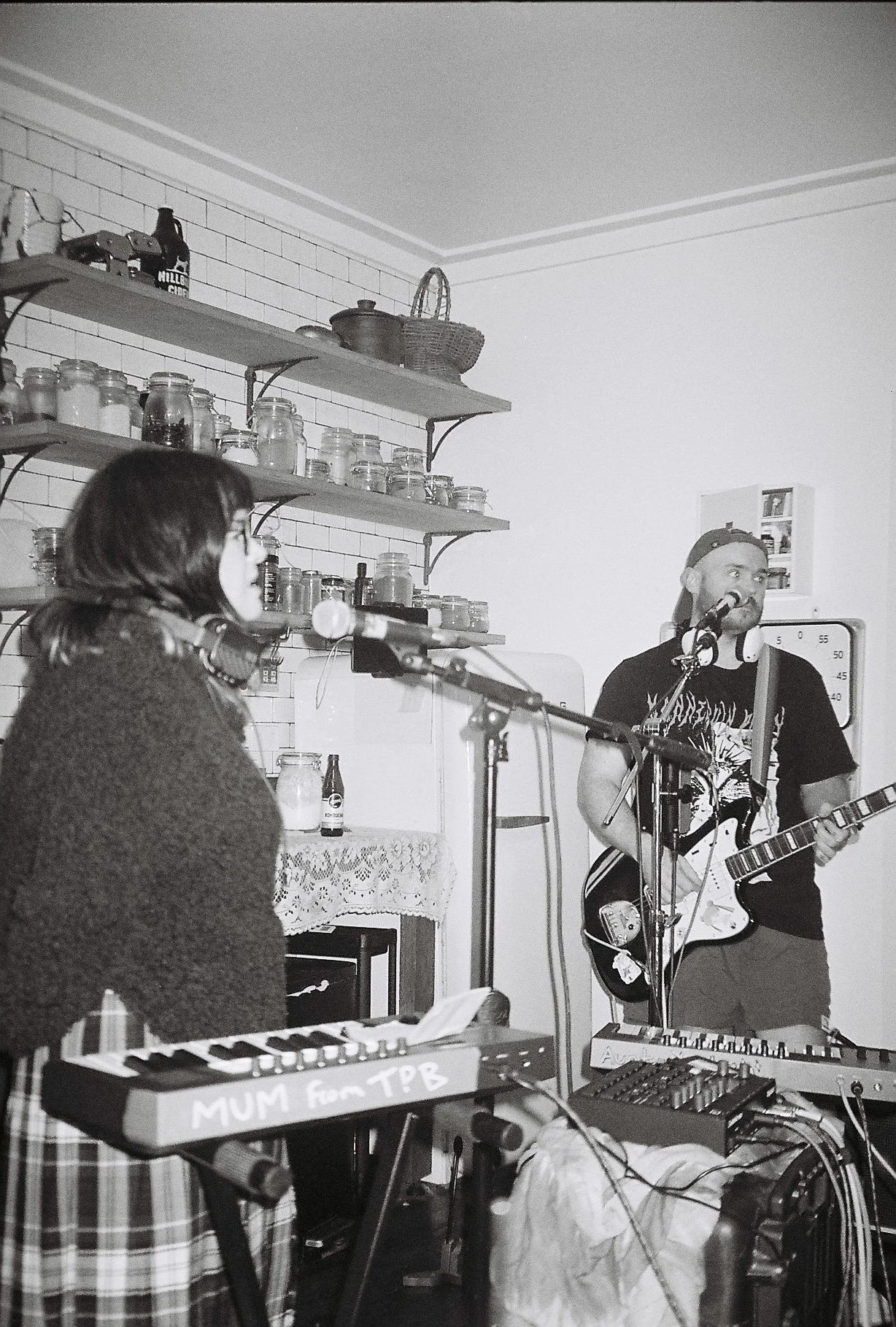 A black and white photo of two musicians performing in a room. A woman with glasses and dark hair is singing into a microphone, while a man with a beard, wearing a cap backwards, is playing an electric guitar. There are musical equipment, shelves with jars, and a laptop in the background.
