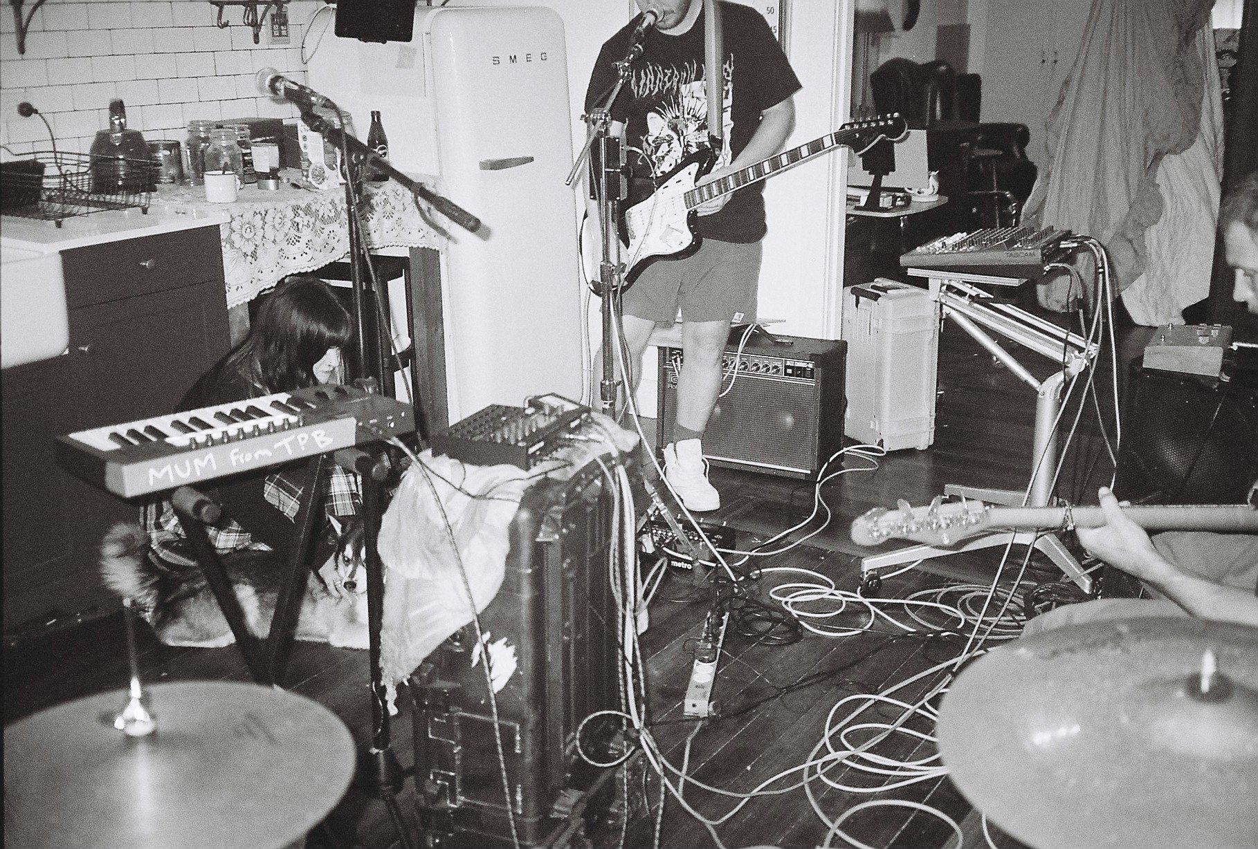 Black and white photo of two children performing music in a cluttered room. One child is playing an electric guitar and the other is sitting on the floor using a keyboard. Signs of instruments, cables, and equipment are visible, with a drummer's cymbal partially seen in the foreground.