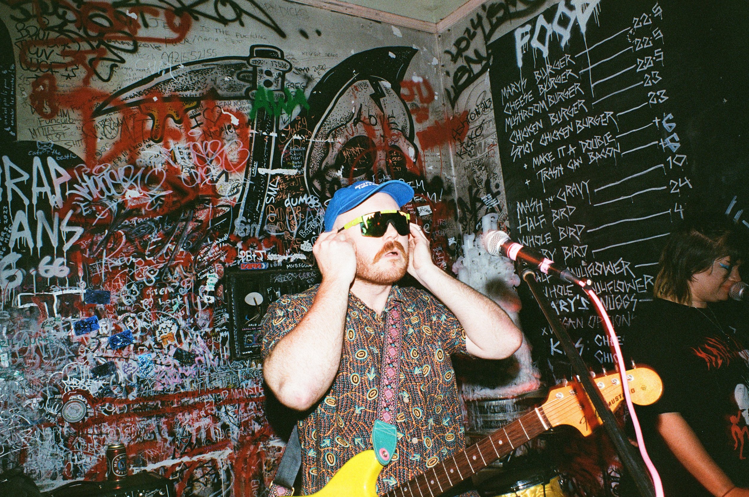 A man wearing sunglasses, a blue cap, and a patterned shirt, holding a guitar in front of a graffiti-covered wall with a menu written on it.
