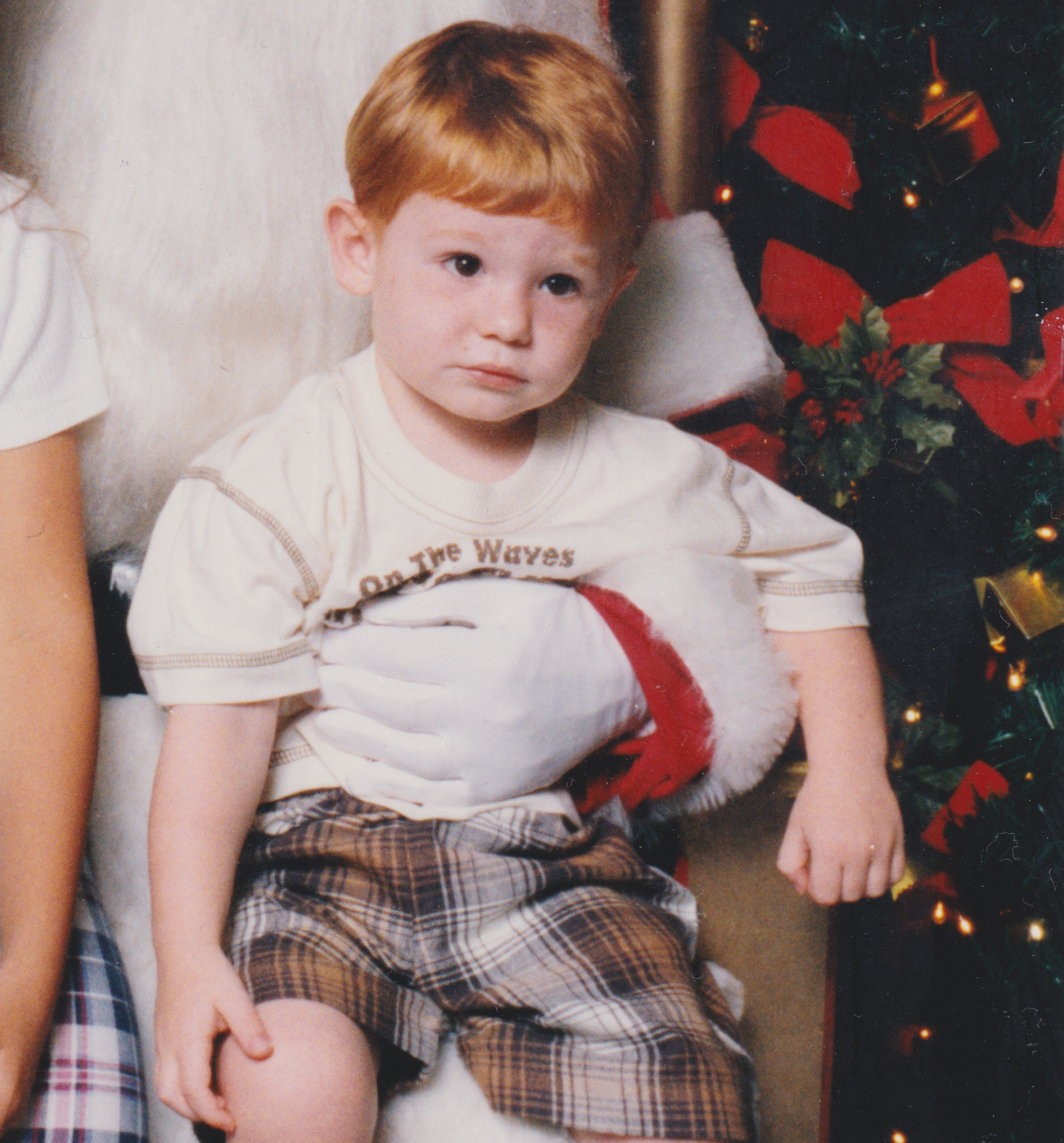 A young boy with red hair sitting next to a Christmas tree decorated with red and gold ribbons and ornaments, wearing a white T-shirt and plaid shorts.