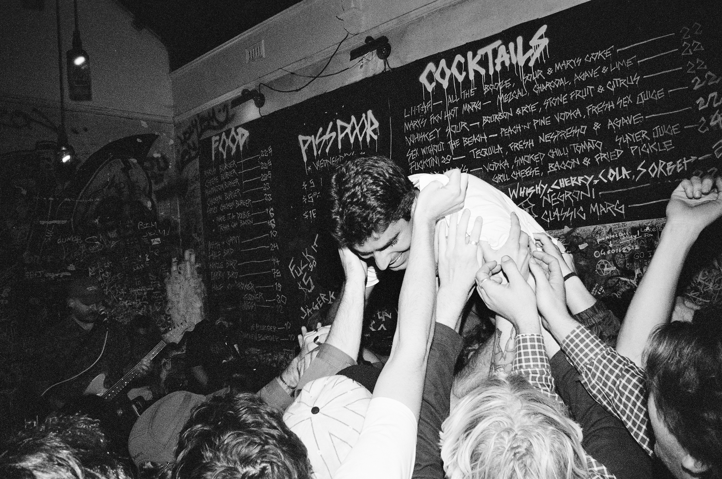 A crowded scene at a party with people holding up a young man, who appears to be crowd surfing, against a blackboard menu of cocktails in the background.