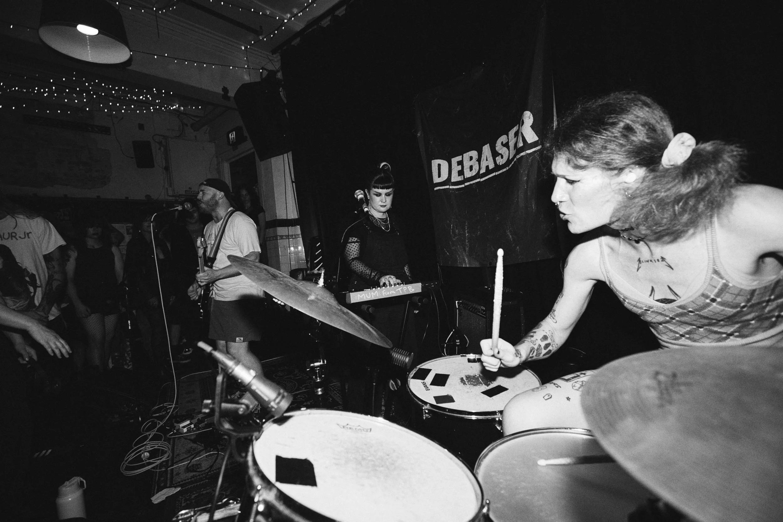 A black and white photo of a band performing in a small, intimate venue. The drummer's kit is in the foreground, with a woman playing drums on the right side of the image. In the background, a man and a woman are visible, with the woman operating a keyboard. The scene is decorated with string lights, and a banner with the word "DEBASER" is hanging on the wall.