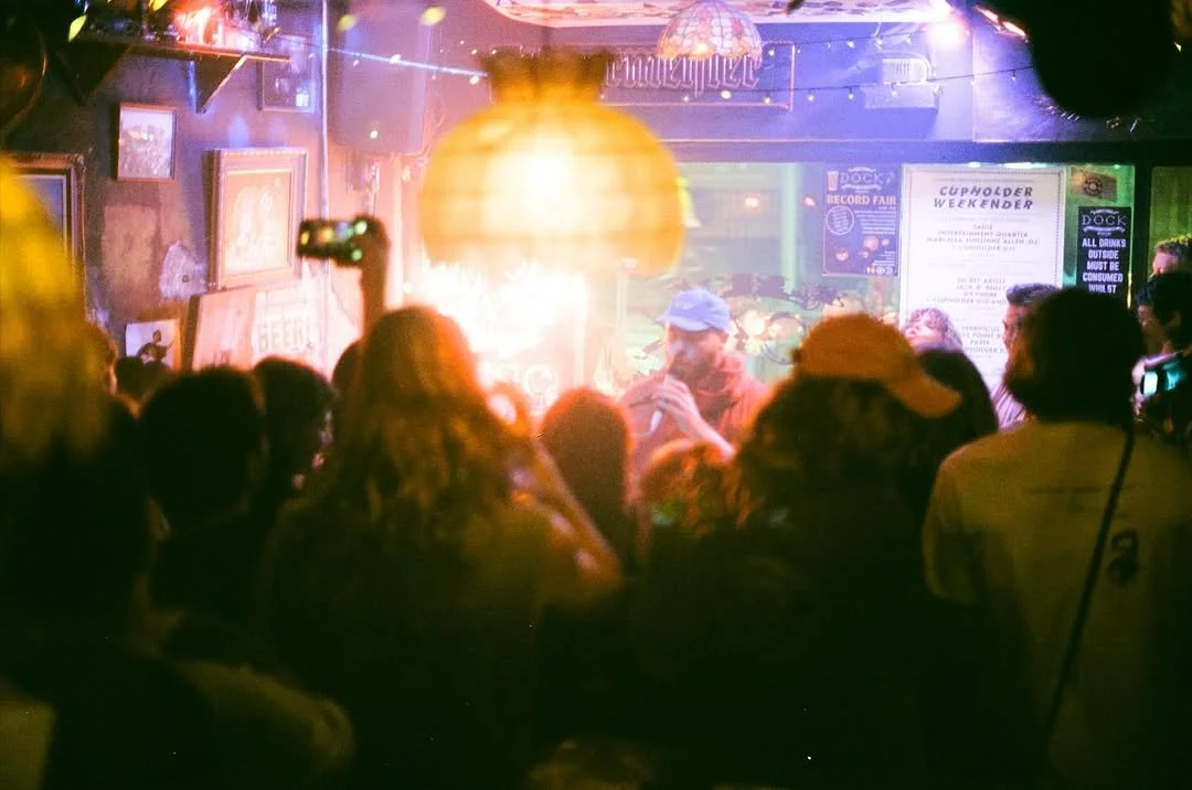 A singer or performer on stage with a microphone, wearing a cap, in a dimly lit bar or club with people gathered and watching, colorful lighting, and posters on the walls.