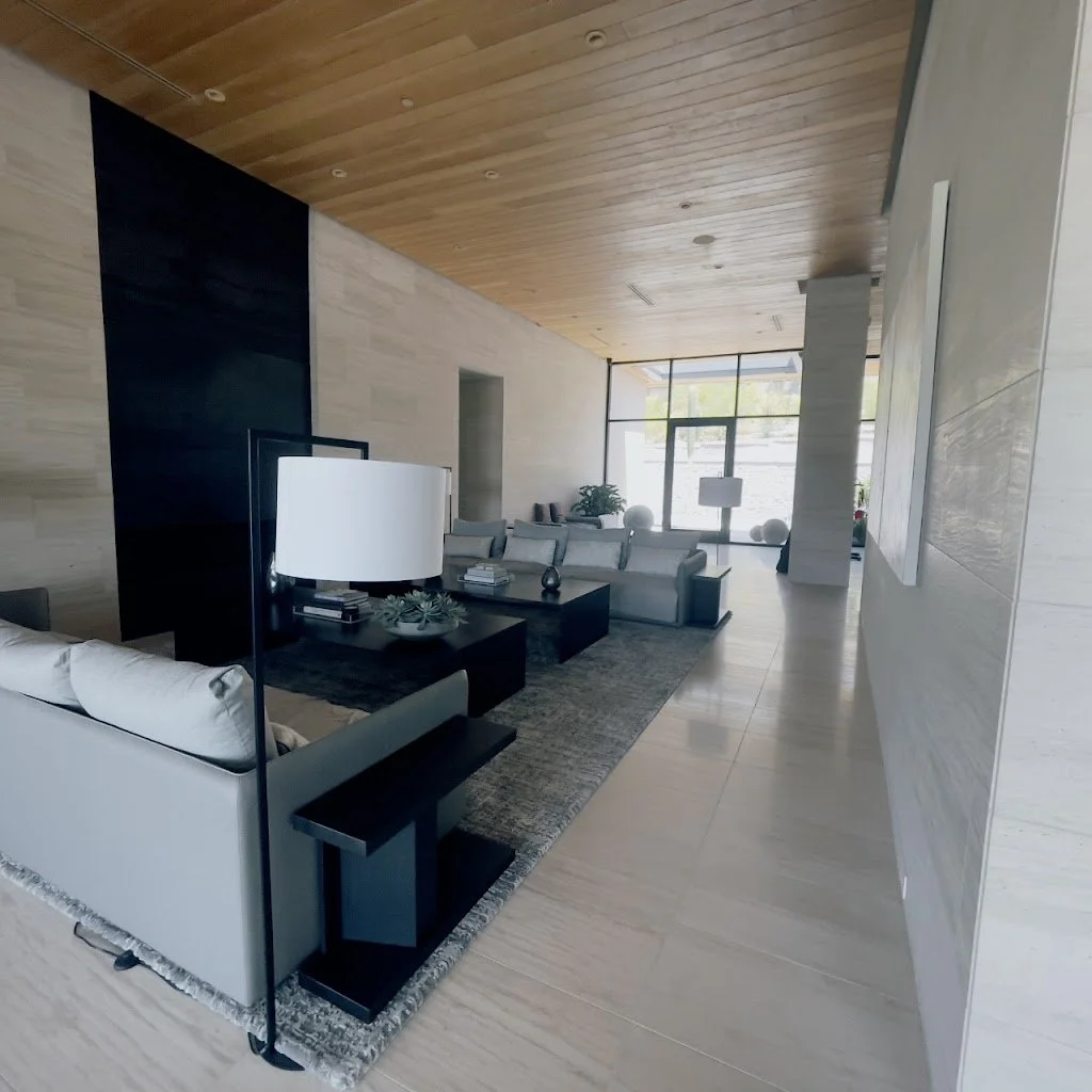 Modern living room with light-colored walls, wooden ceiling, and large windows in the background. Contains white sofas, black tables, and decorative lamps, with a gray area rug on the floor.