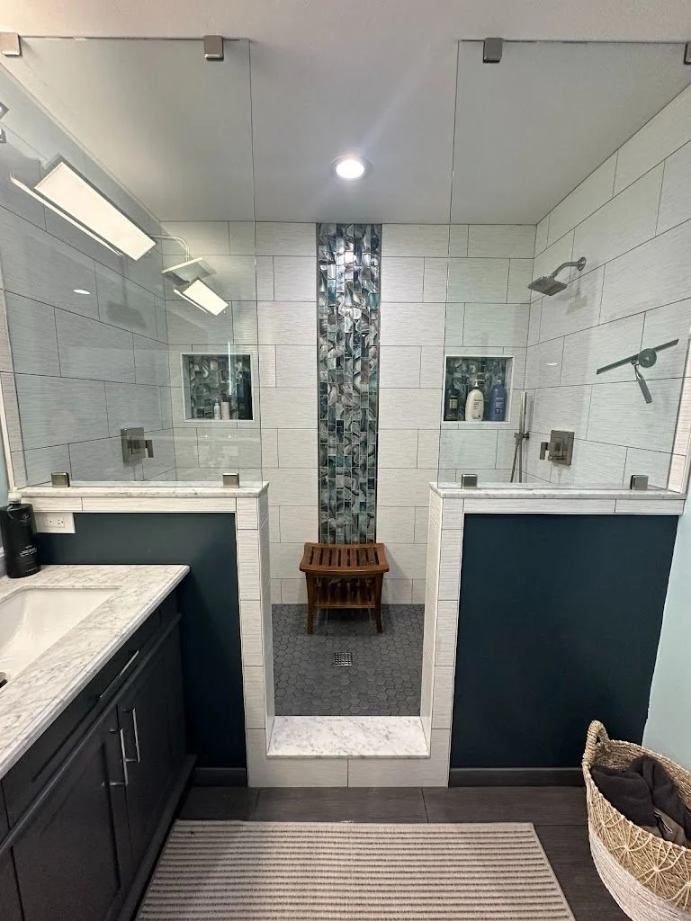 View of a modern bathroom with a dual shower area separated by glass with dark blue walls, white subway tiles, built-in shower shelves, a small wooden bench, and mosaic tile accents.