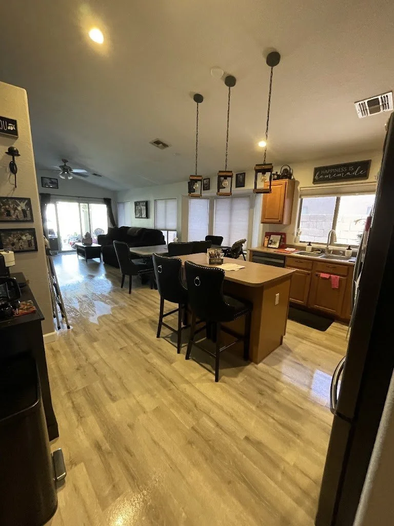 Open kitchen and living room with wooden flooring, a kitchen island with three black chairs, and a dining area with a table and chairs. In the background, there are large windows and sliding glass doors leading outside. The ceiling has three hanging pendant lights.