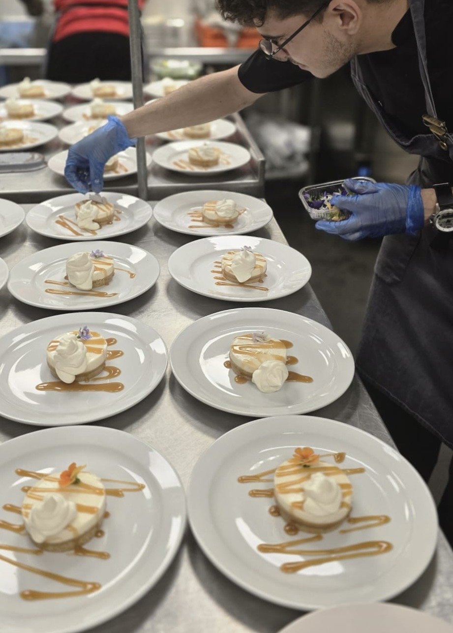 A chef in a commercial kitchen preparing plated desserts with a caramel sauce, whipped cream, and edible flowers on white plates.