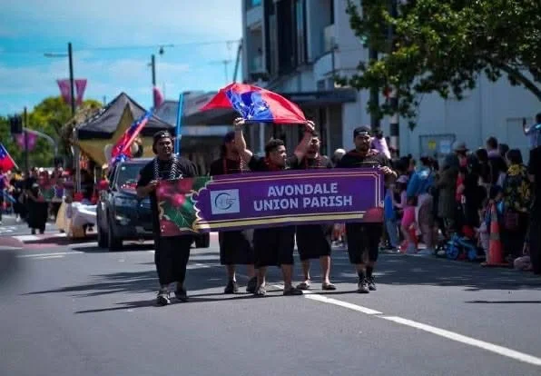 People walking in a parade holding a purple banner that reads "Avondale Union Parish." One person holds a colorful umbrella, and there are spectators along the sidewalk.