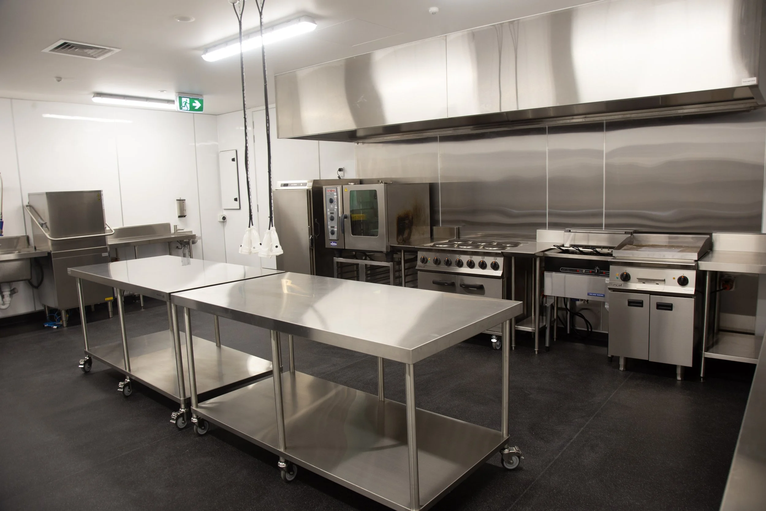 Empty commercial kitchen with stainless steel tables, appliances, and ventilation hood, illuminated by overhead fluorescent lights.