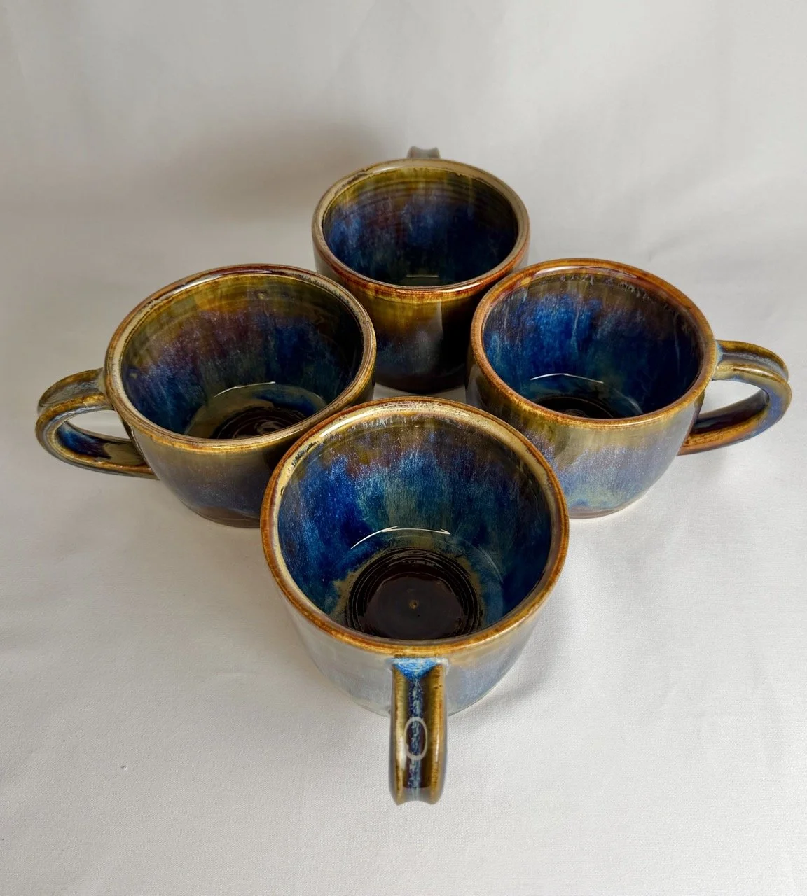 Four ceramic mugs with a multicolored glaze pattern, positioned with their openings facing upward on a white surface.