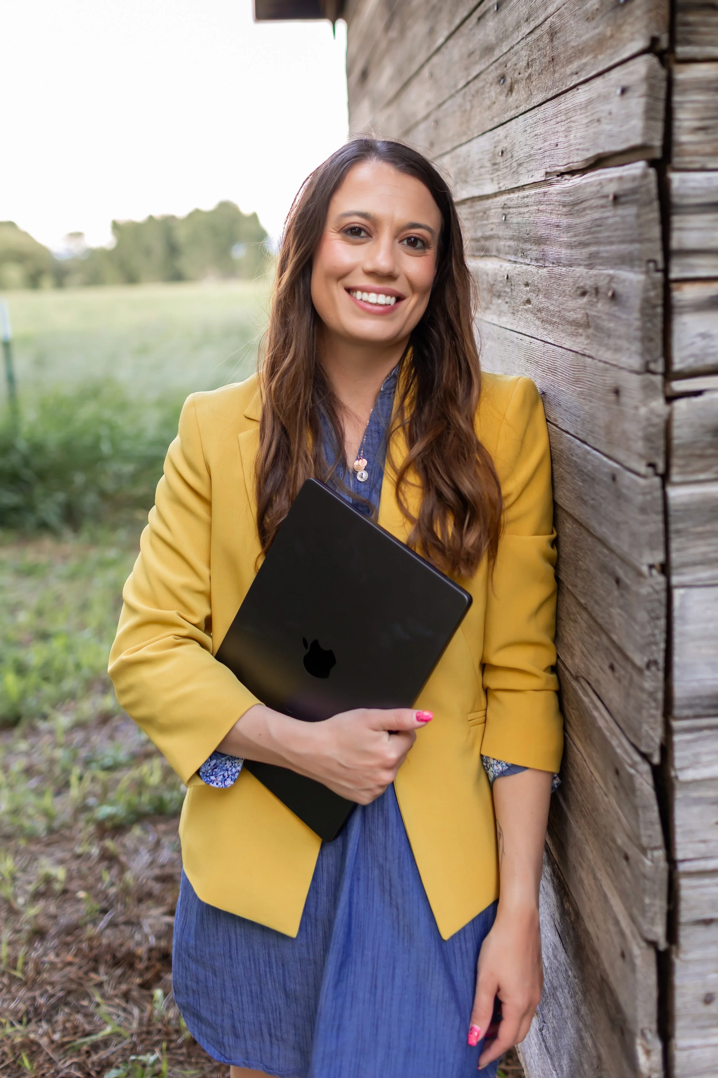 A woman with long brown hair, wearing a yellow blazer and blue dress, standing outdoors, holding a black Apple laptop, smiling, leaning against a wooden barn wall.