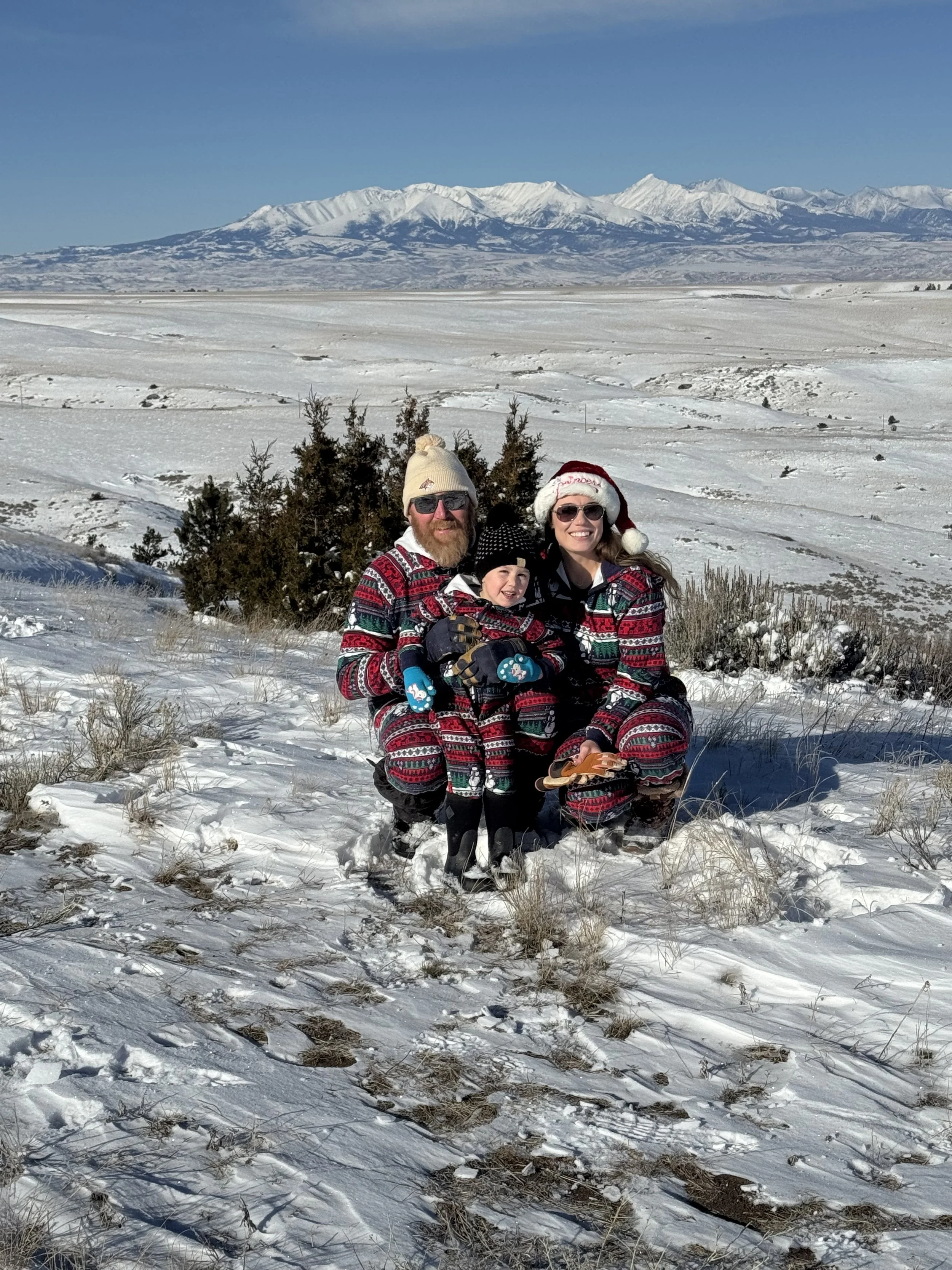Family of three dressed in matching Christmas sweaters and winter hats, kneeling in snow on a hillside with snow-covered mountains in the background.