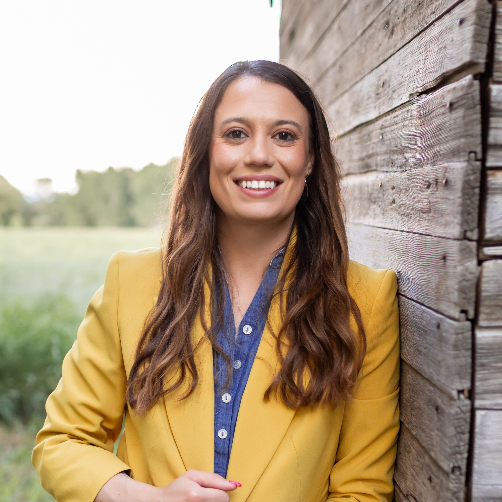A woman with long brown hair smiling, wearing a yellow blazer over a blue button-up shirt, standing outdoors near a wooden wall with a grassy field and trees in the background.