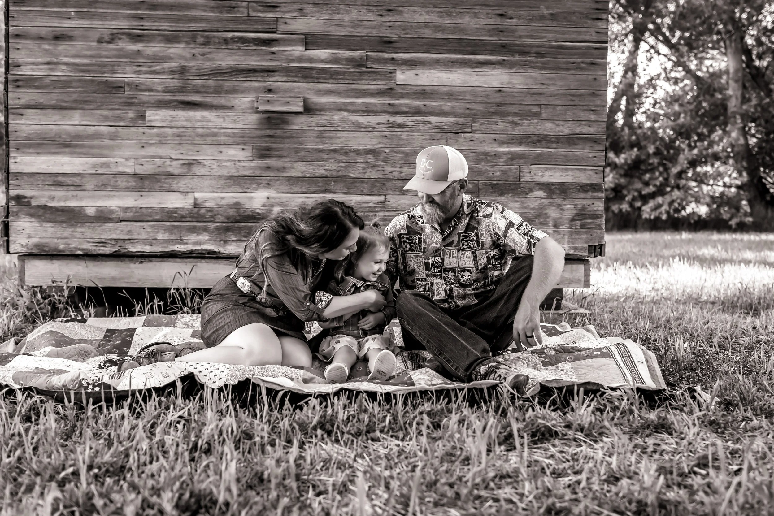A family of three, consisting of a mother, father, and young child, sitting together on a blanket outside next to a wooden building. The family appears to be enjoying a moment of laughter and closeness.