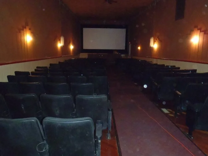 Empty movie theater with rows of black chairs, a large screen at the front, and dim wall lights along the sides.