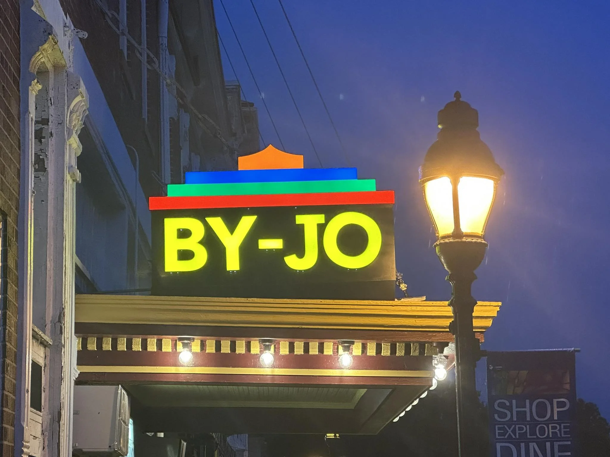A neon sign displaying the word "BY-JO" with colorful layers above it, located on a building sidewalk at night, near a lit street lamp.