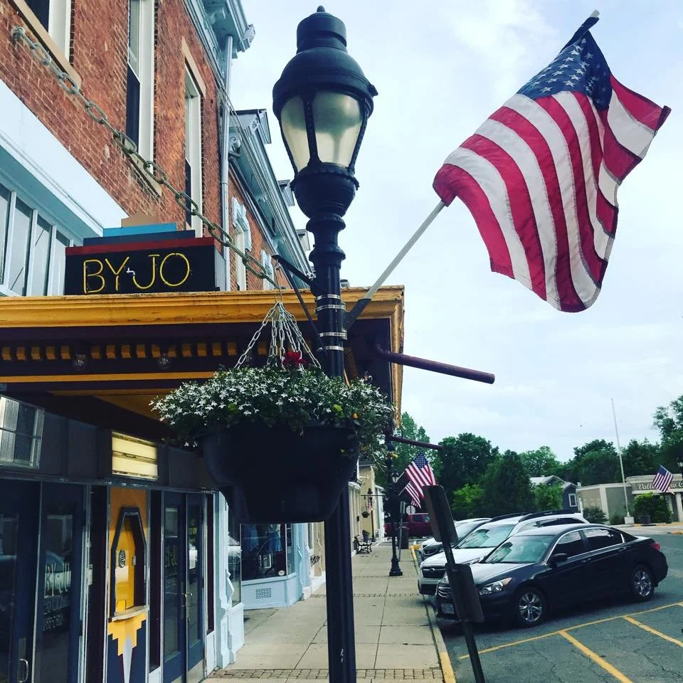 A street view showing a brick building with a sign that says BYJO, a black street lamp with hanging flower baskets, and American flags mounted on the lamp and in the parking lot. Cars are parked along the street.