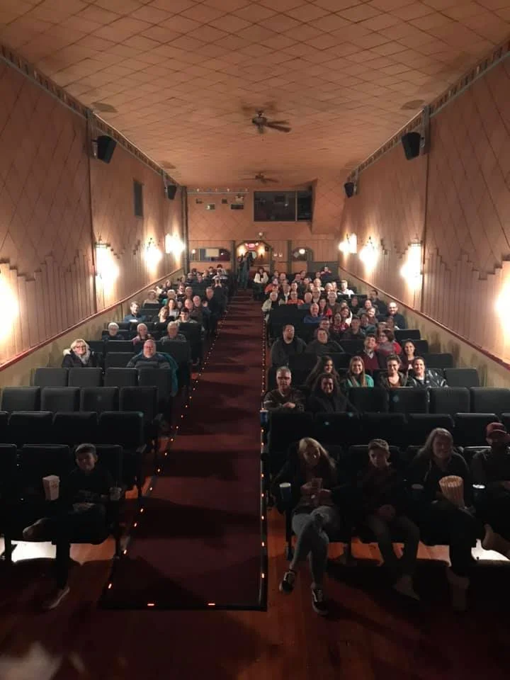 Audience seated in a small theater or cinema, waiting for a movie or performance to start, with a red carpet aisle down the center and warm lighting