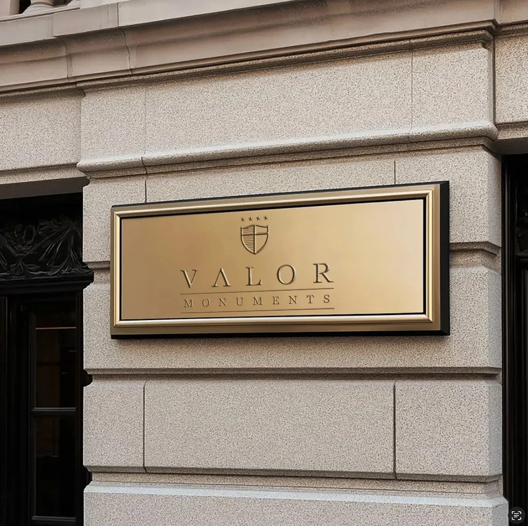 Gold and black sign reading 'Valor Monuments' with a crest above it, mounted on a beige brick building wall.
