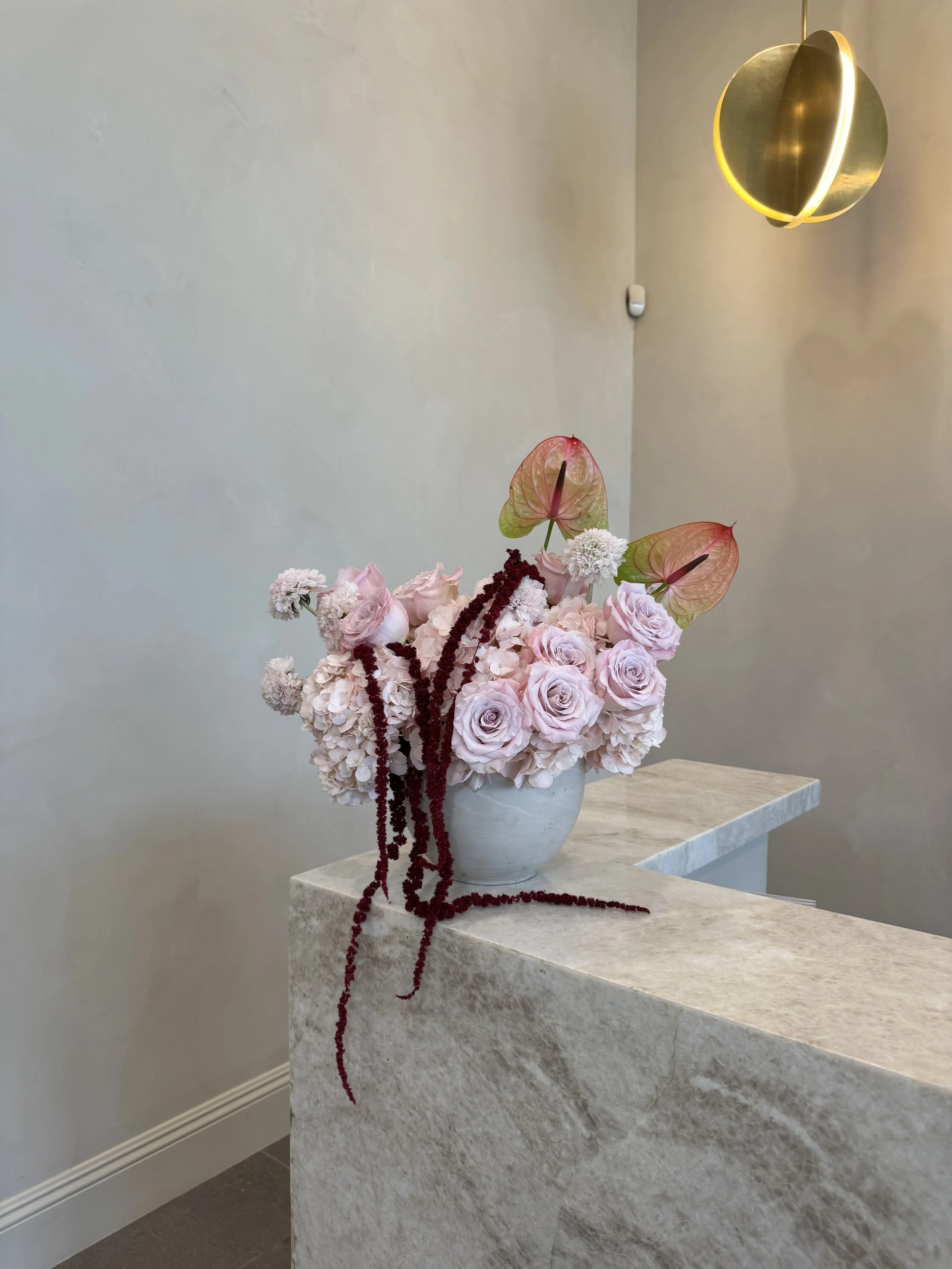 A floral arrangement with pink roses, white carnations, and other flowers in a white vase on a marble countertop, with a modern gold ceiling light overhead.