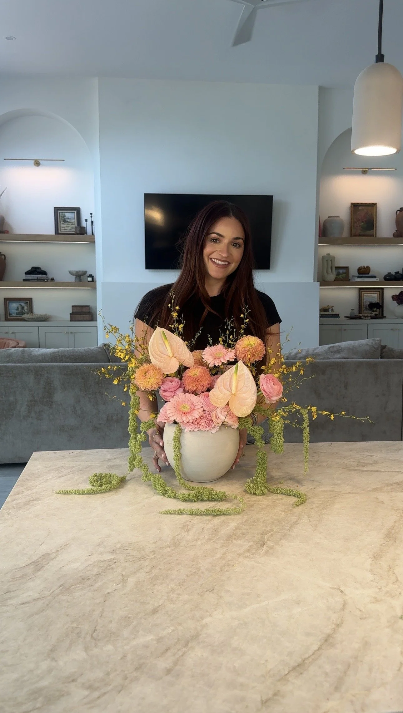 Woman smiling and holding a flower arrangement in a beige vase on a light-colored table in a modern living room.