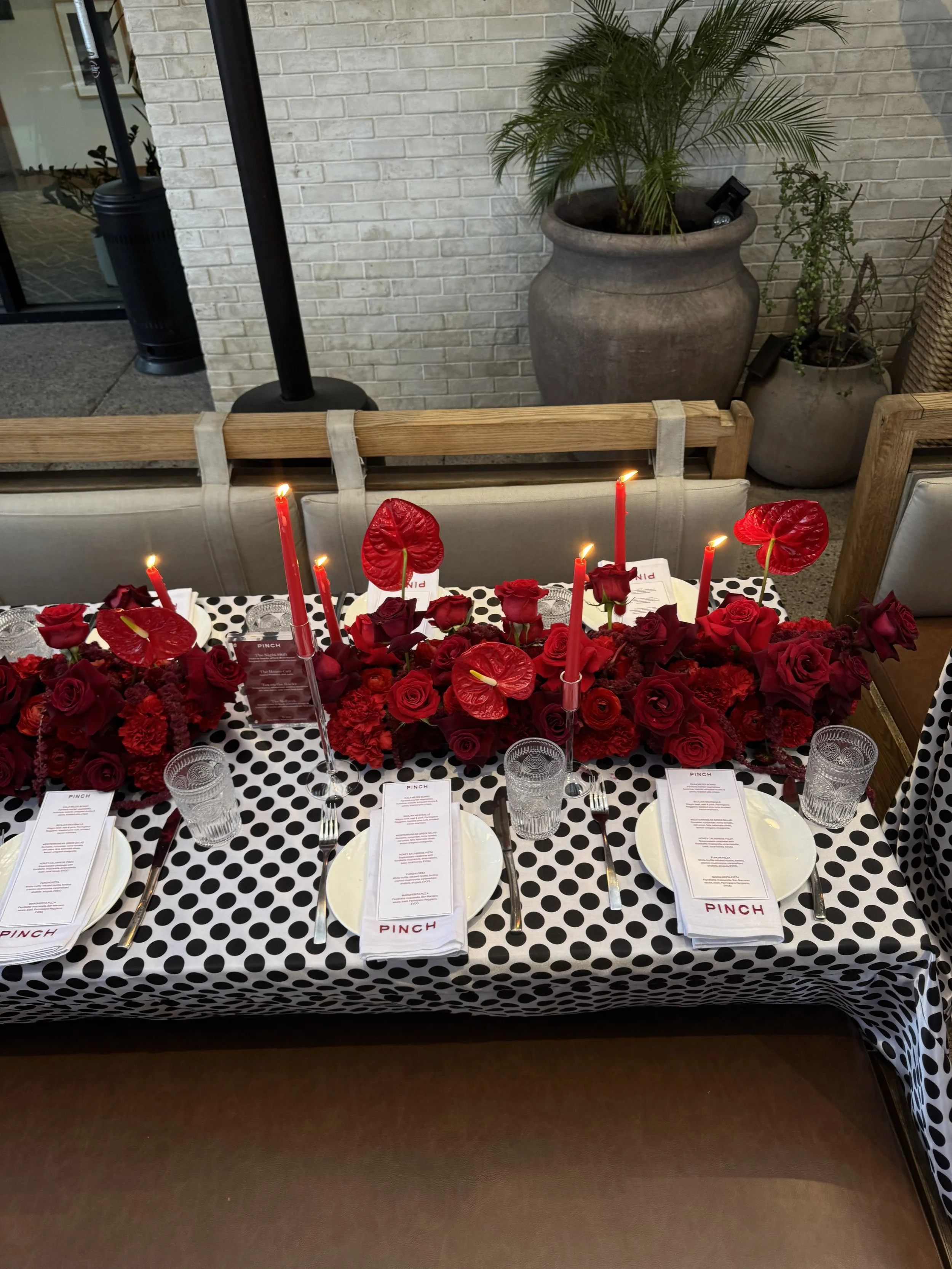 A table decorated with a black and white polka dot tablecloth, red candles, and red flowers, set for a meal with plates, glasses, and menu cards labeled 'PINCH' at each place setting. Behind the table are two large pots with indoor plants, and a bric