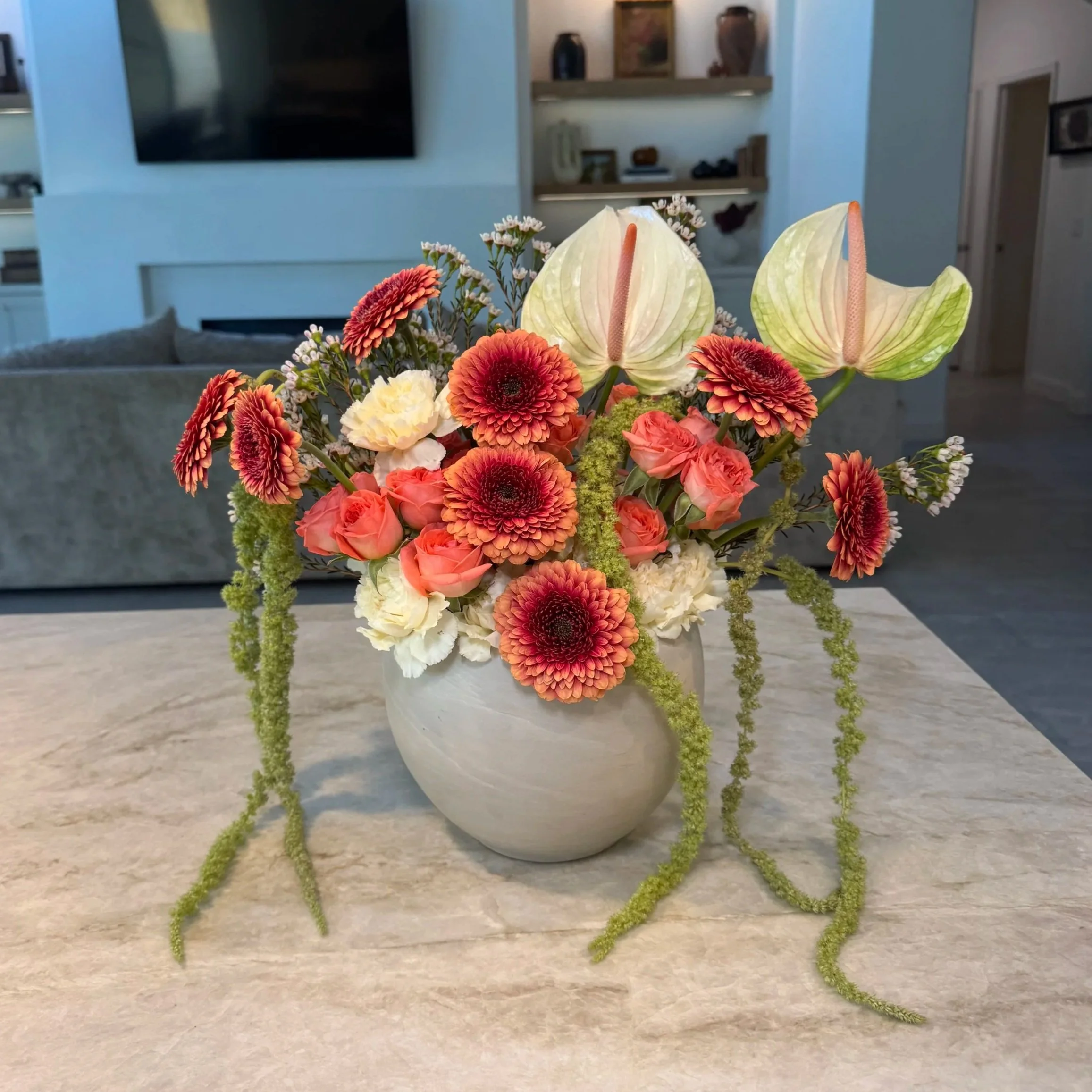 A bouquet of various flowers including lilies, chrysanthemums, and other wildflowers in a white ceramic vase placed on a beige surface in a living room with a television and decorative items on shelves in the background.