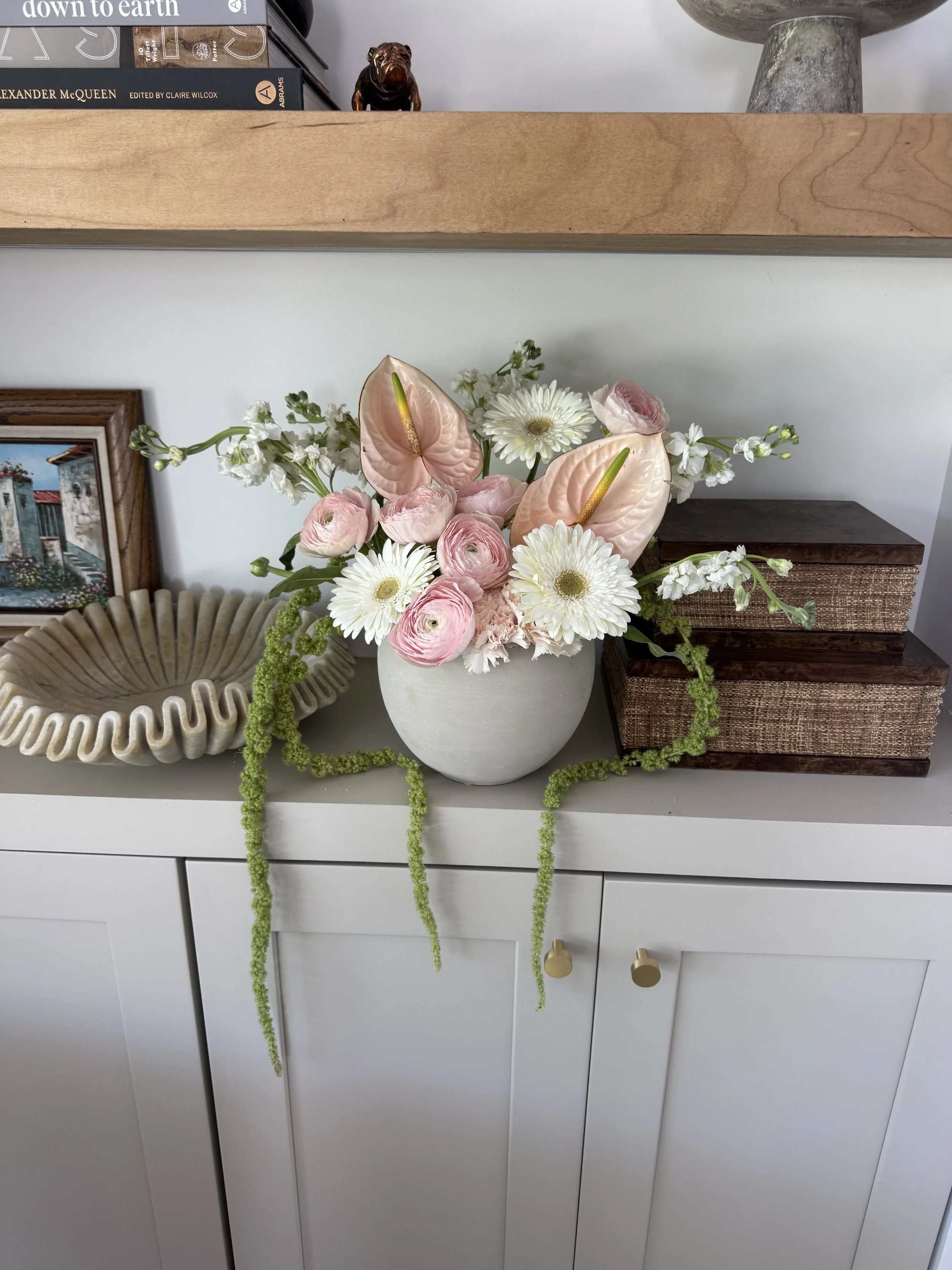 A white vase filled with pink and white flowers, including anthuriums and daisies, placed on a white dresser with a wooden box to the right and decorative objects to the left. A framed picture and a rattan bowl are also on the dresser.