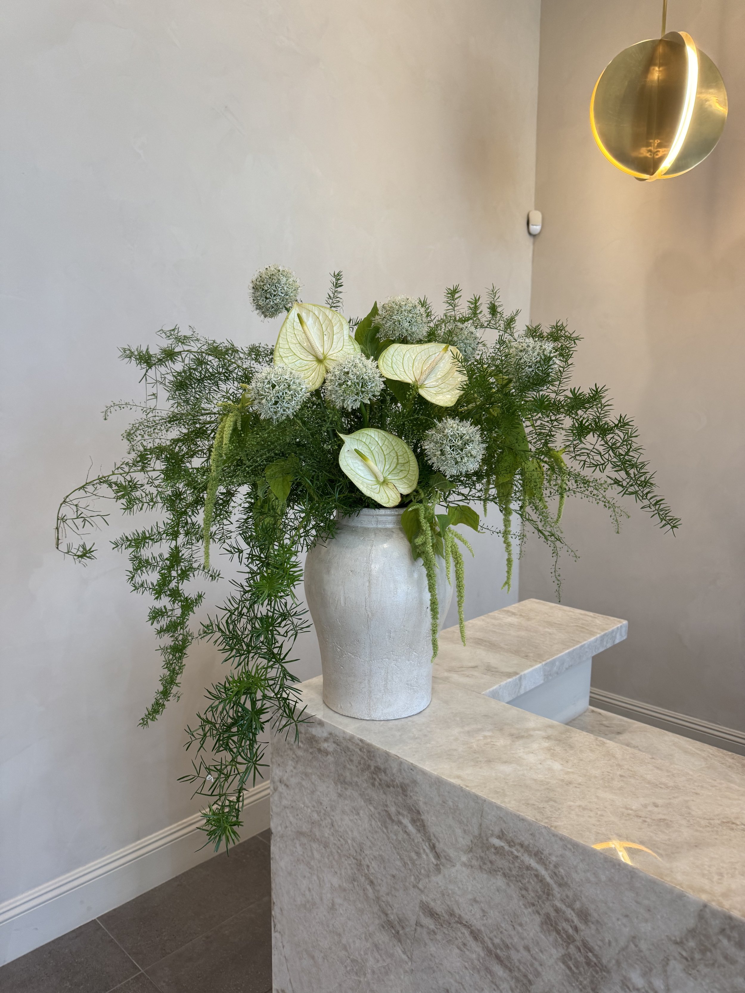 A large floral arrangement with white and green flowers and lush green foliage in a tall white vase, situated on a stone counter in a modern interior with a plain wall and contemporary pendant lighting.