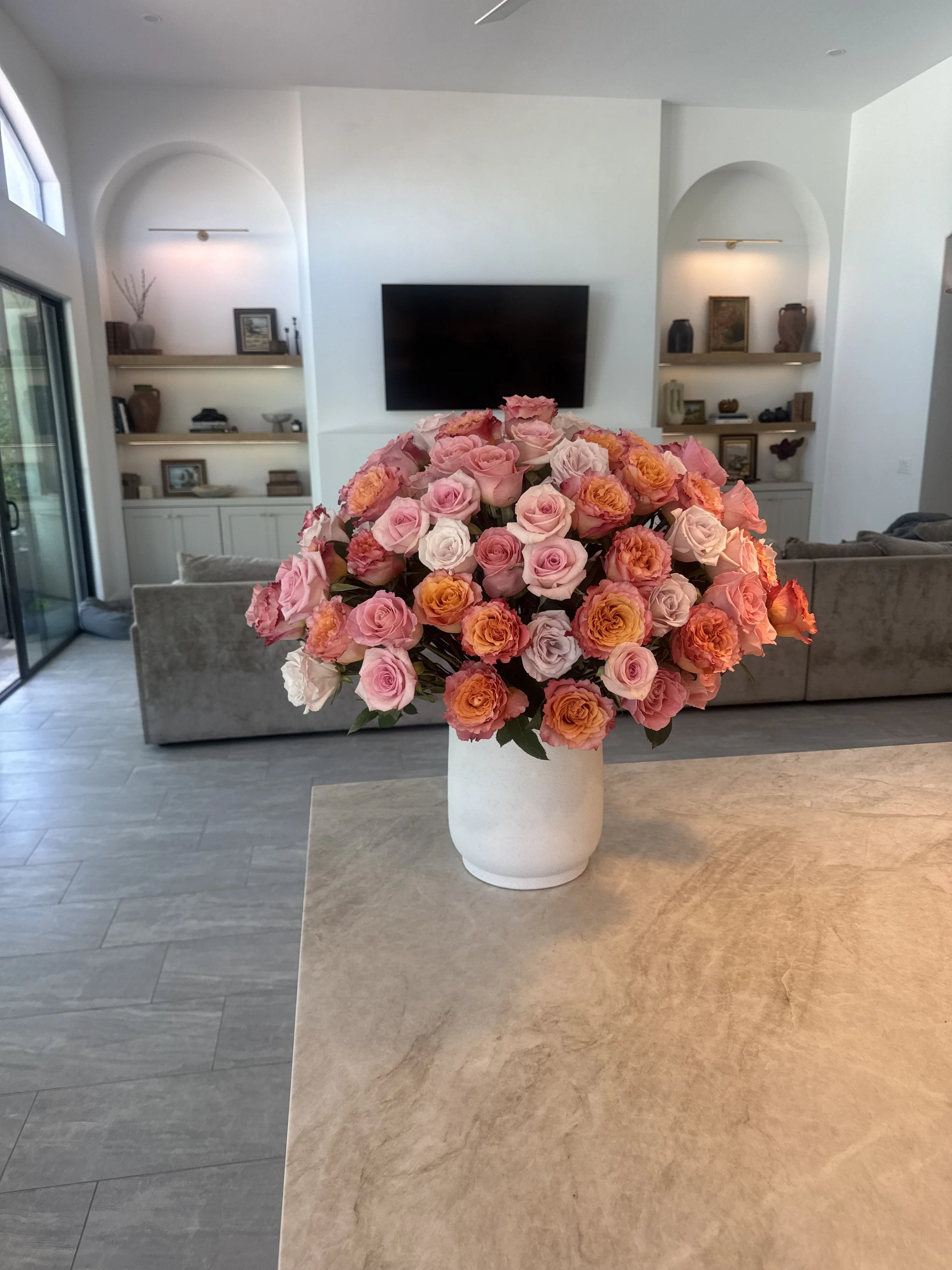 A large bouquet of pink and orange roses in a white vase on a light-colored kitchen or dining table.