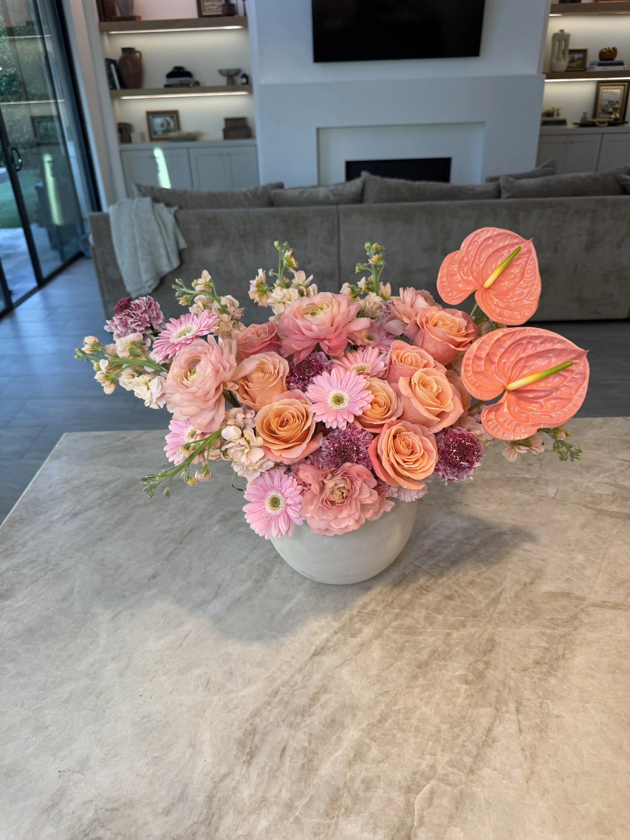 A beige vase filled with pink, orange, and purple flowers, placed on a light-colored marble table in a modern living room with a gray sofa, white built-in shelves, and a TV.