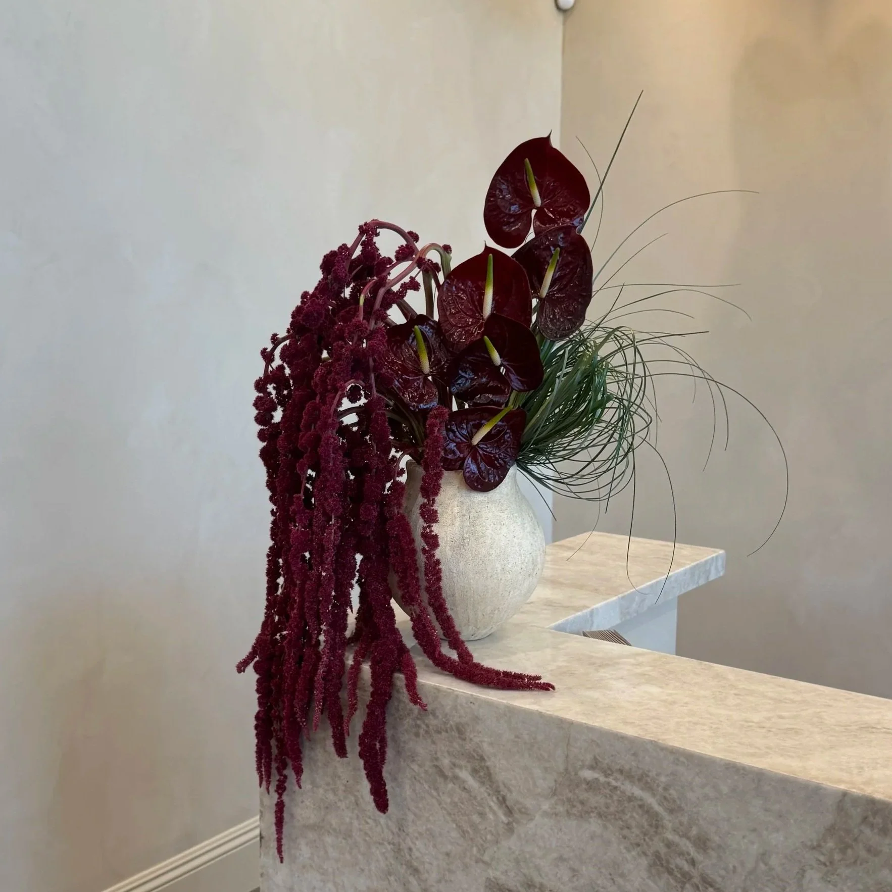 A large decorative flower arrangement in a white vase, featuring deep red anthuriums, pink amaranth, and long slender green leaves, placed on a marble surface against a plain beige wall.