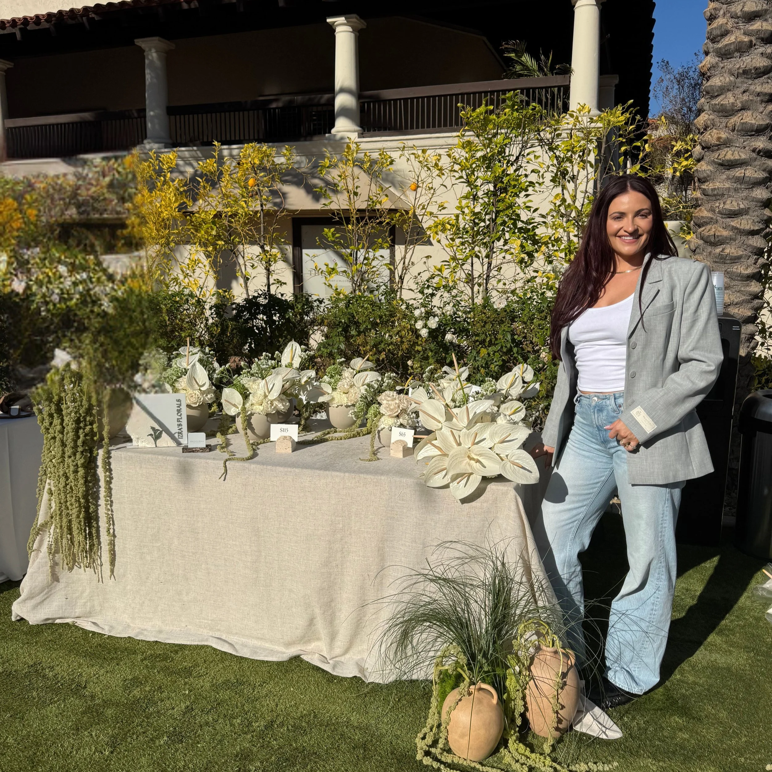 A woman with long dark hair, wearing a light gray blazer and white shirt, standing outdoors in front of a table with white floral arrangements and decorative stones. The background features a house with a balcony, columns, and a garden with various plants and trees.