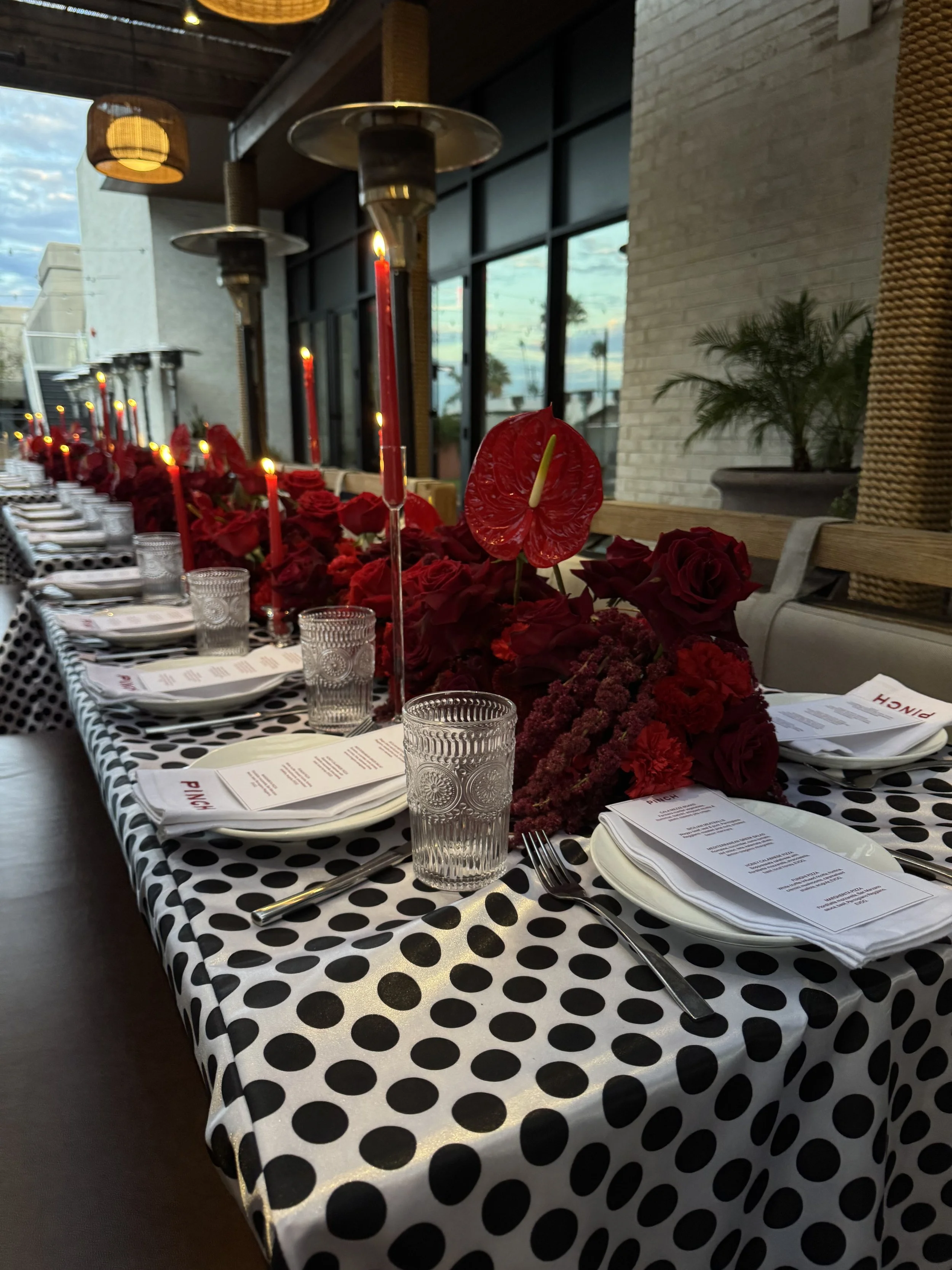 A long dining table covered with a black and white polka dot tablecloth, decorated with red roses and tall red candles, set with glassware, plates, napkins, and utensils in a warmly lit indoor space with large windows and view of the sky and palm tre