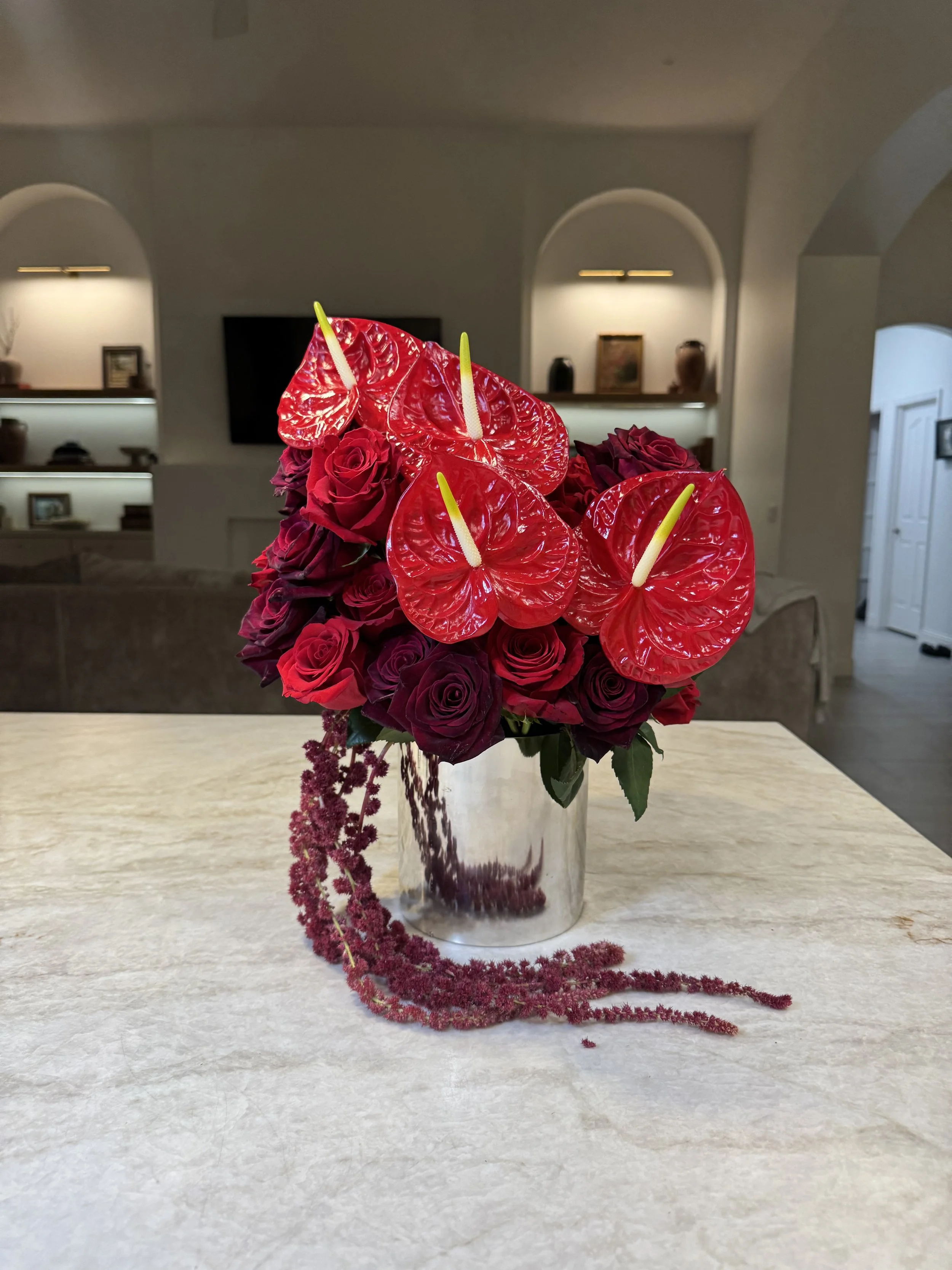 A bouquet of red roses and red anthuriums with yellow pistils in a marble-patterned vase on a white tabletop.