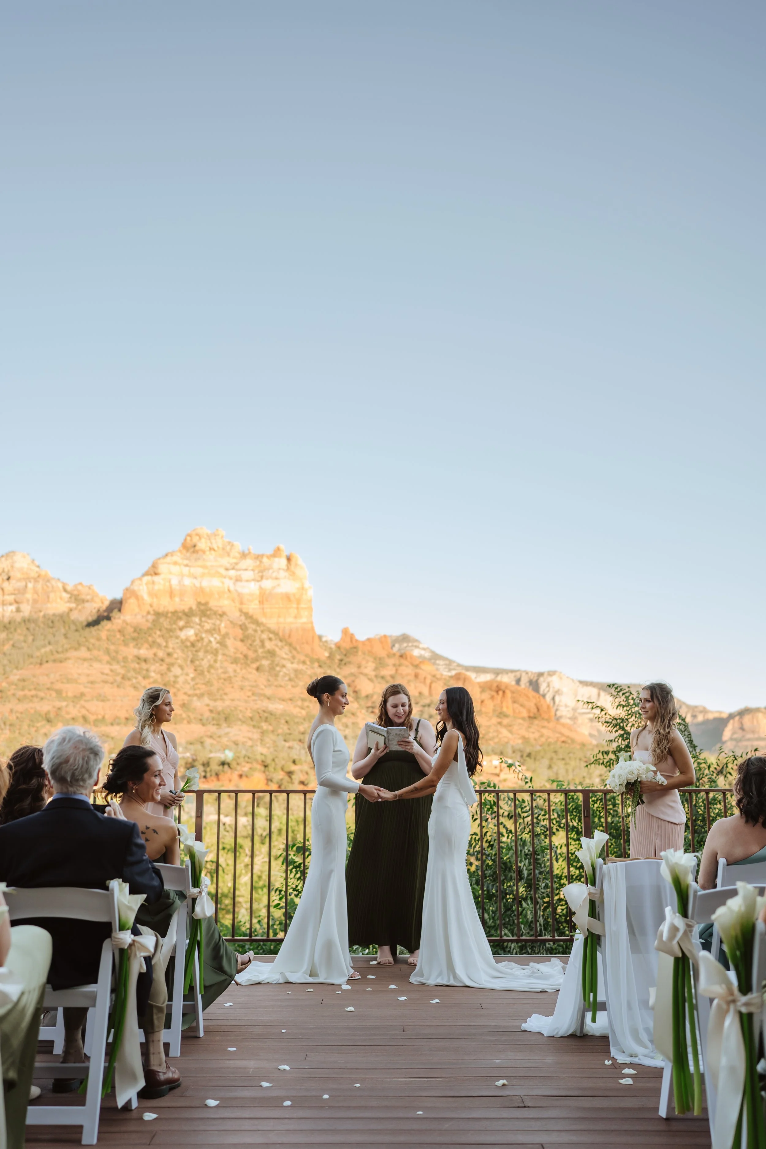 A wedding ceremony with two brides holding hands, standing in front of an officiant, with guests seated on either side. The ceremony takes place outdoors on a wooden deck with scenic mountains in the background and decorated with white flowers and ri