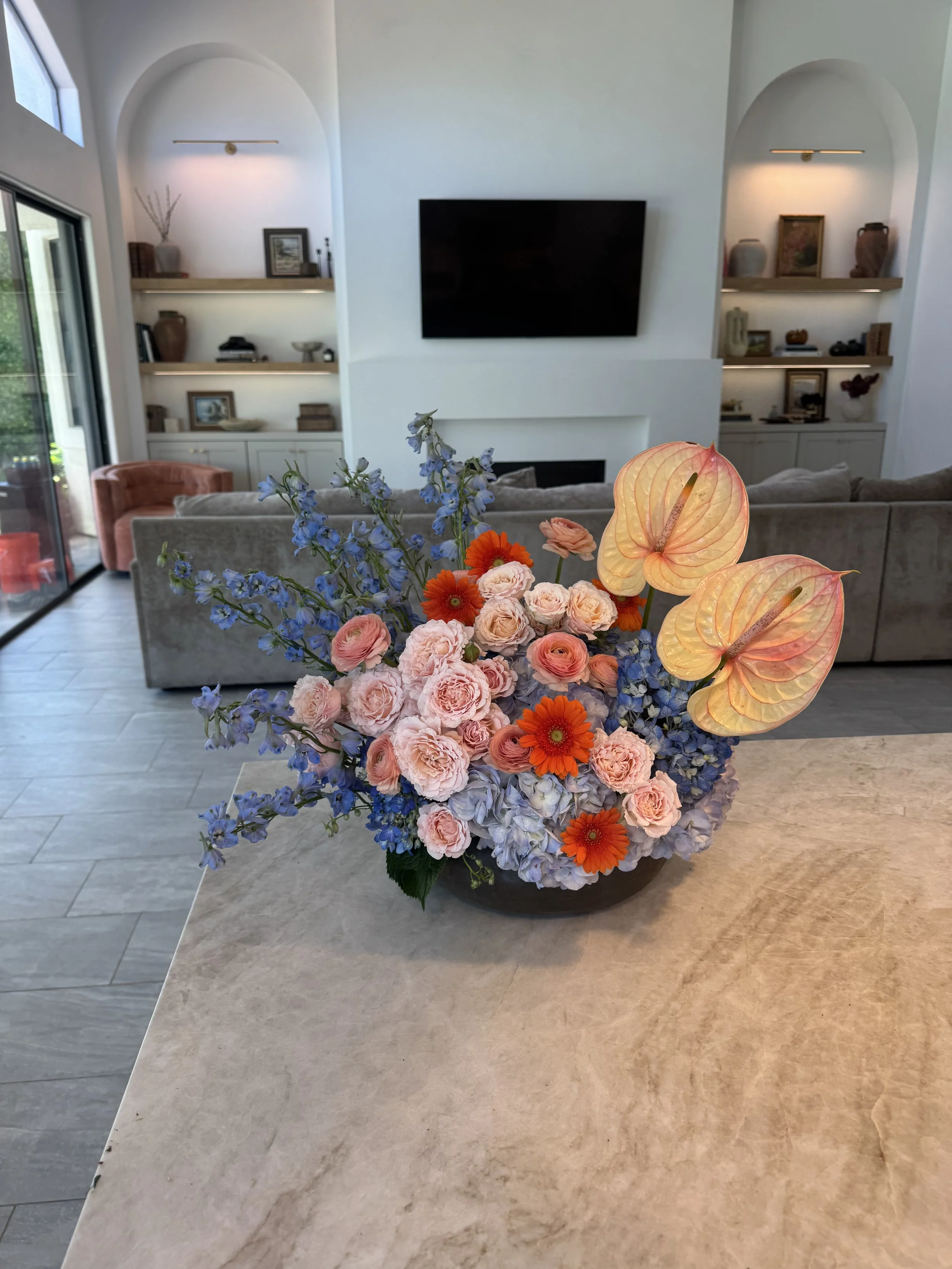 A floral arrangement with pink roses, orange daisies, blue delphiniums, and peach-colored anthuriums on a light-colored marble countertop in a modern living room with built-in shelves, a wall-mounted TV, and a sitting area.