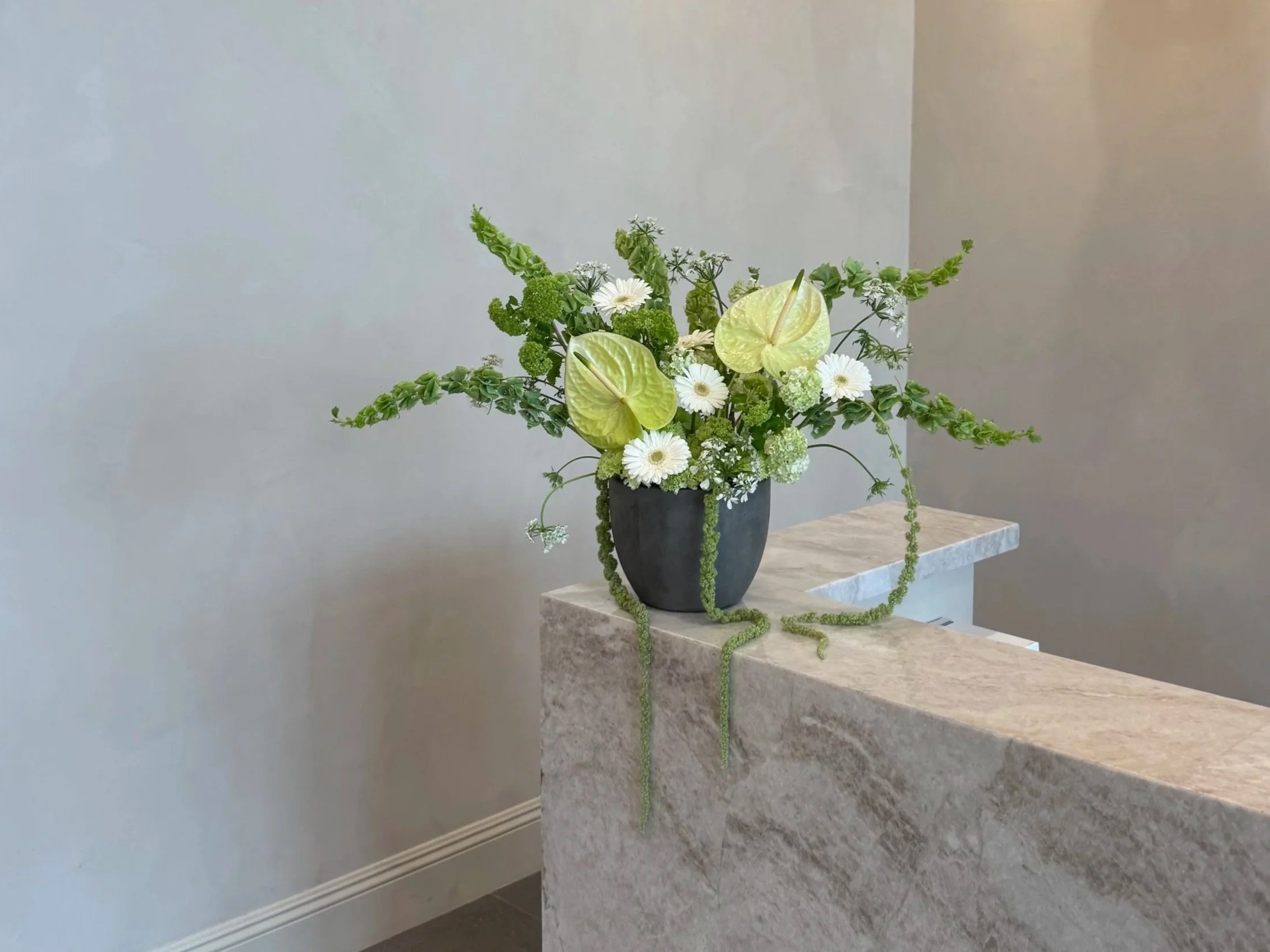A floral arrangement in a black vase placed on a marble countertop. The bouquet contains white daisies, green anthuriums, green roses, and various green foliage, set against a plain, light-colored wall.