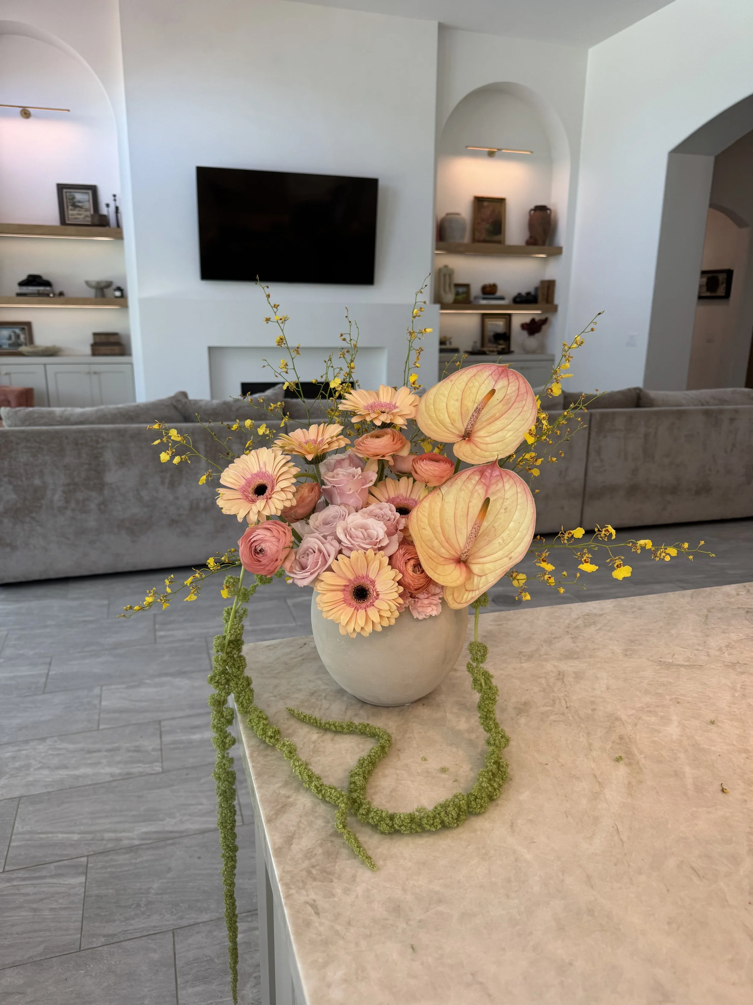 A white ceramic vase filled with pink, peach, and cream-colored flowers, including gerbera daisies, roses, and anthuriums, with trailing green moss on a beige marble table in a living room.